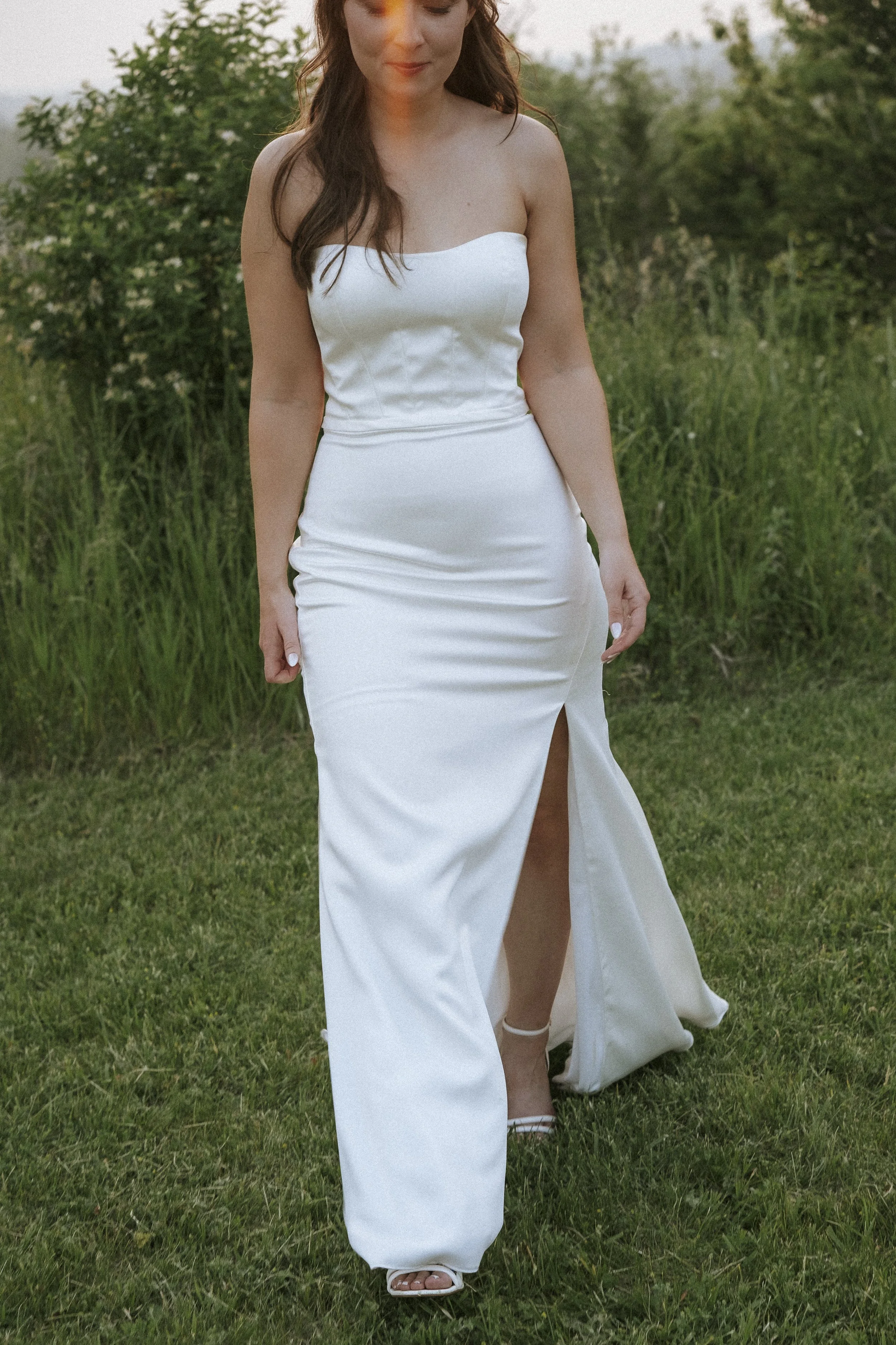A woman in a white strapless dress walking on grass outdoors with greenery in the background.