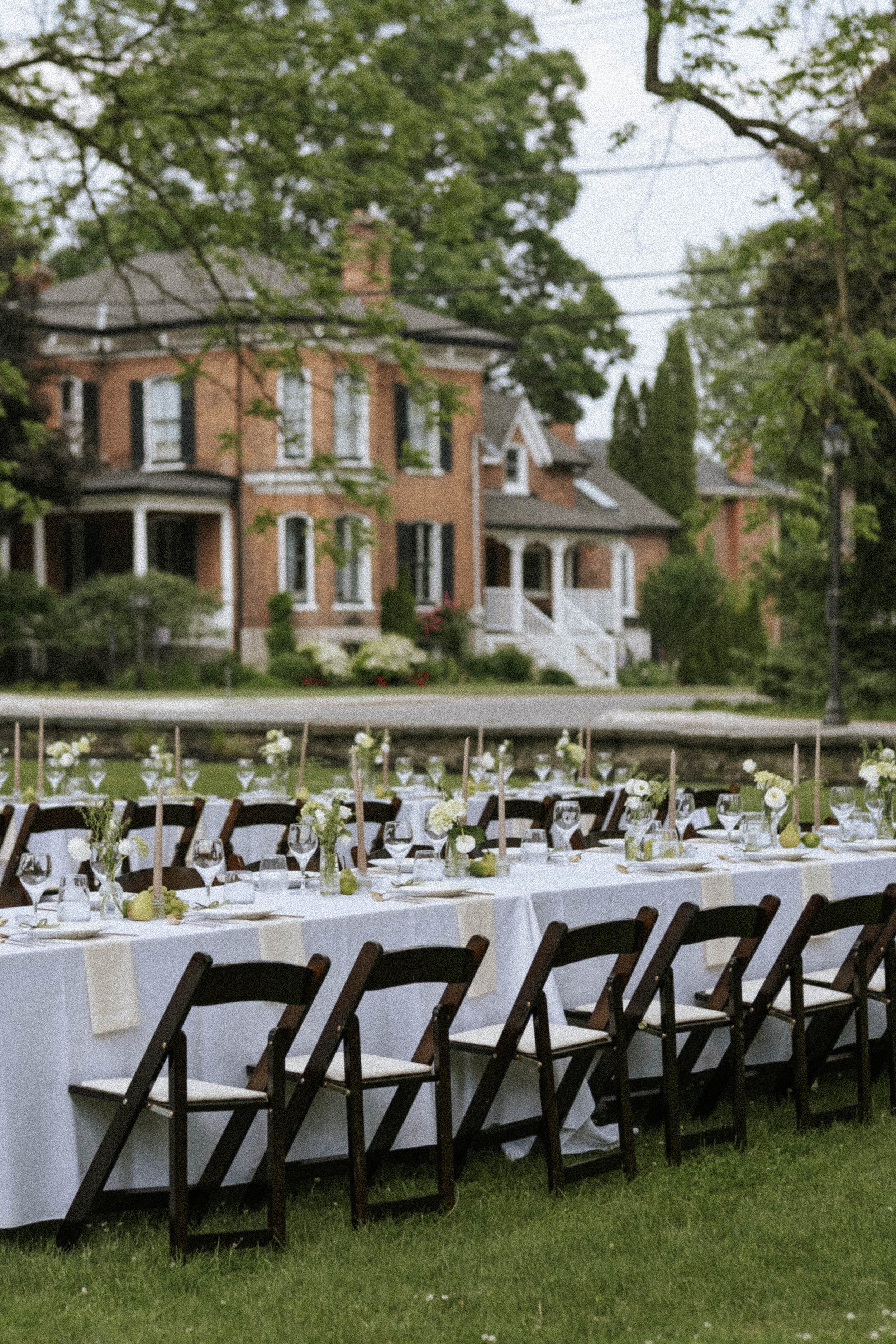 An outdoor dining setup with a long table covered in a white tablecloth, decorated with flowers and glassware, set up on a lawn in front of a large brick house with lush trees.