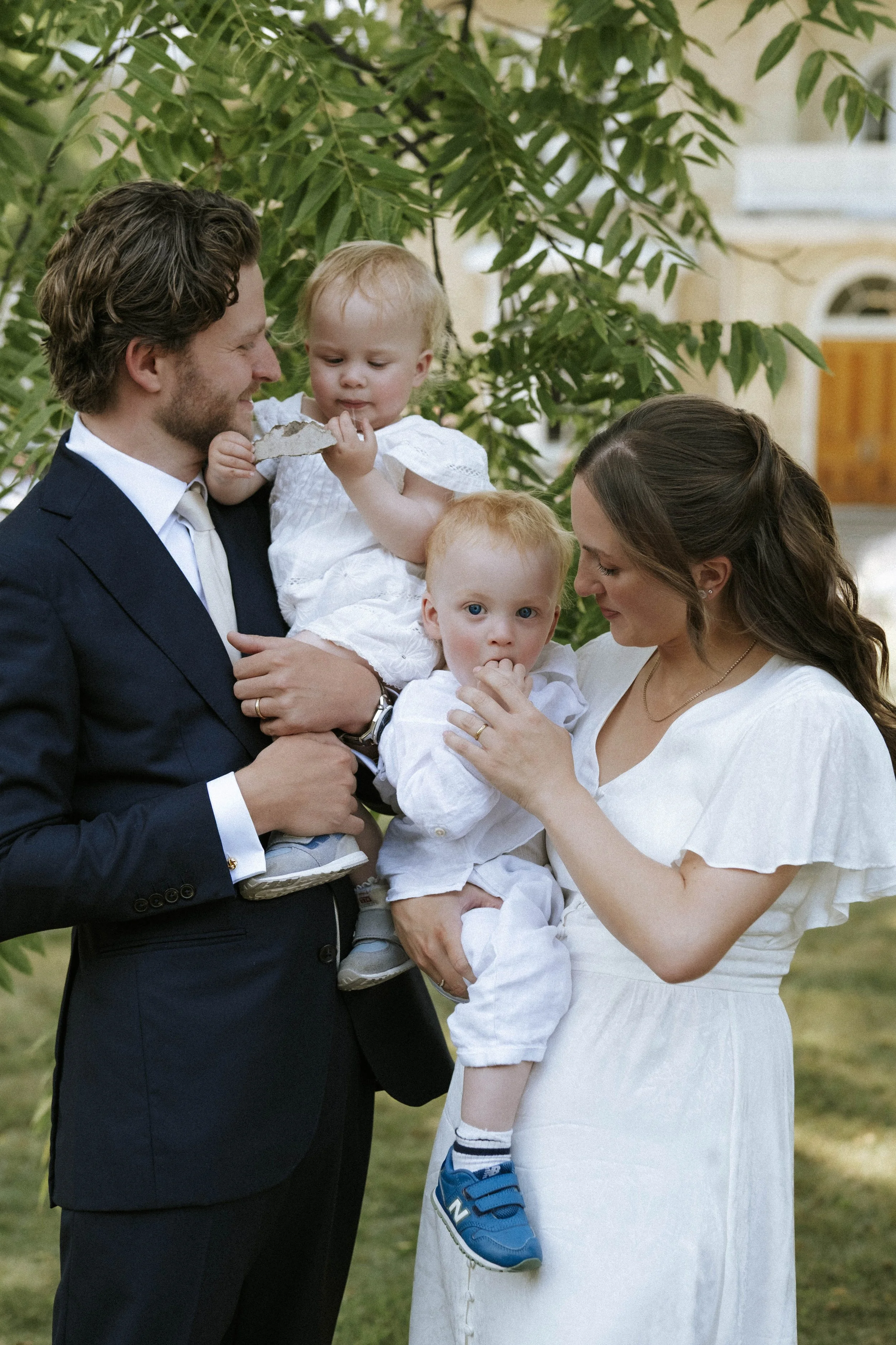 A family of four outside, with a man in a suit, a woman in a white dress, and two young children, one holding a rock and the other eating a hand. They are standing in front of greenery and a house.