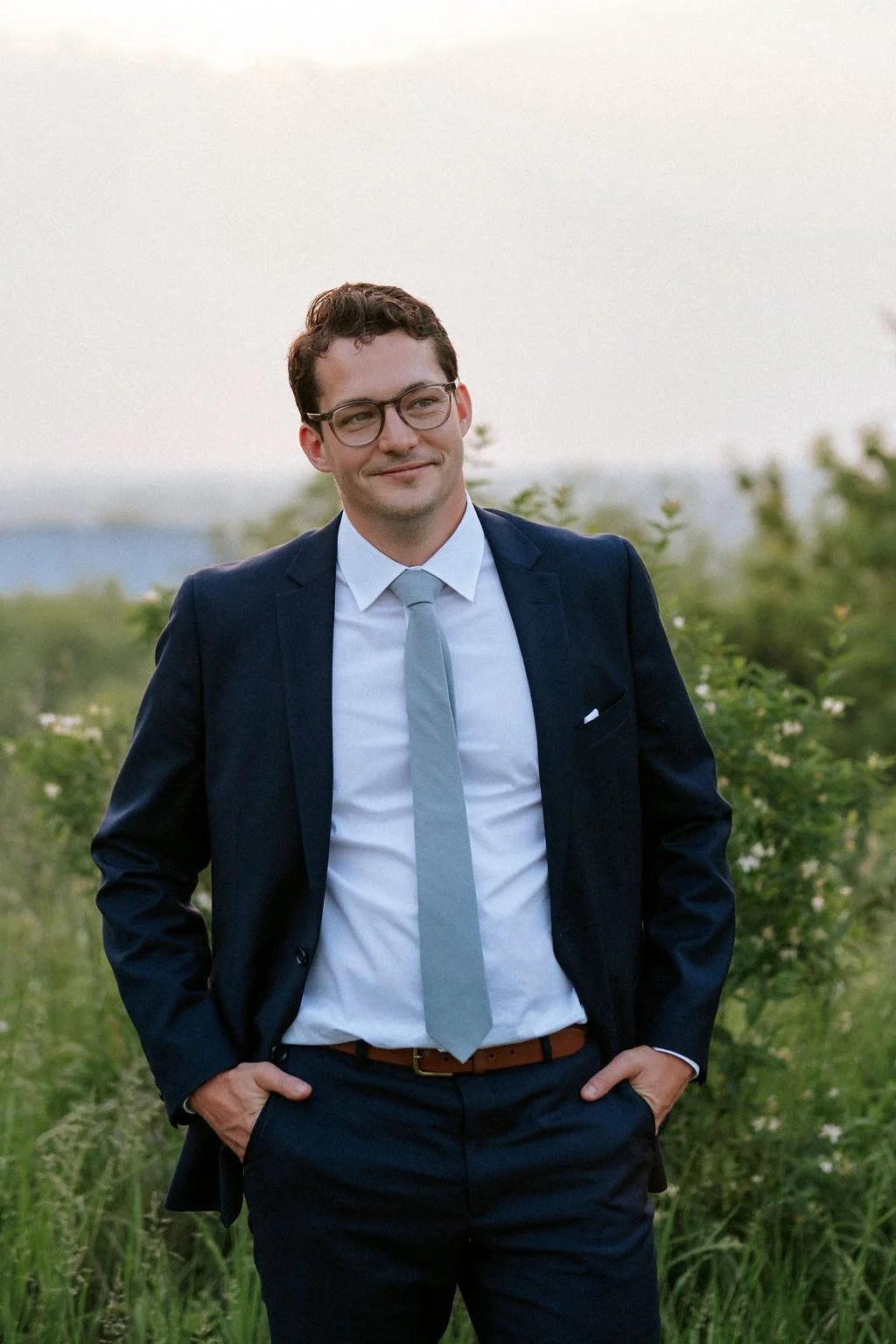 A young man wearing glasses, a dark suit, a white shirt, and a light gray tie, standing outdoors with green foliage and a distant body of water in the background, smiling gently.