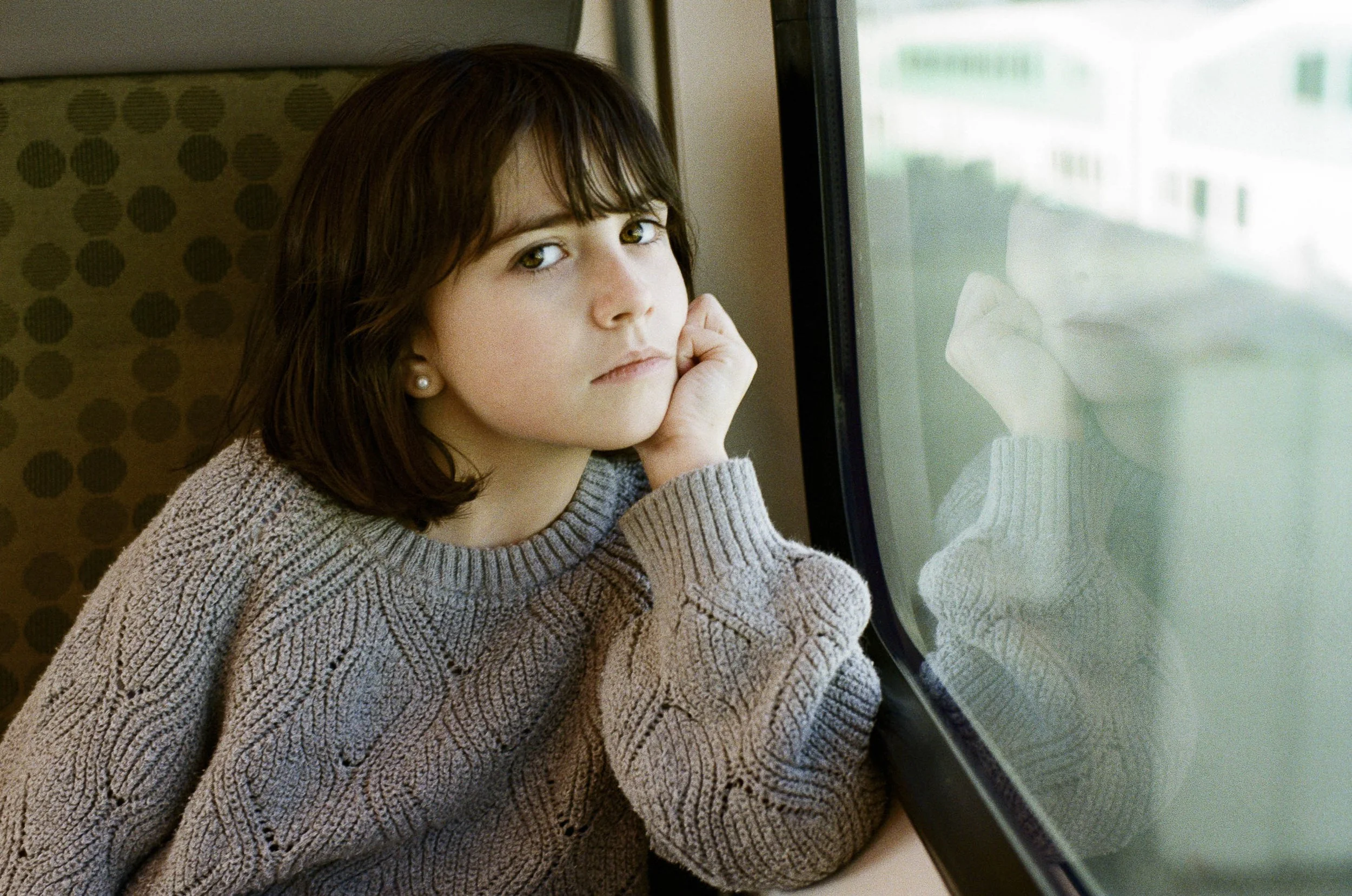 A young girl with dark brown hair and light skin looks out of a window with a pensive expression, resting her face on her hand, wearing a knitted gray sweater.