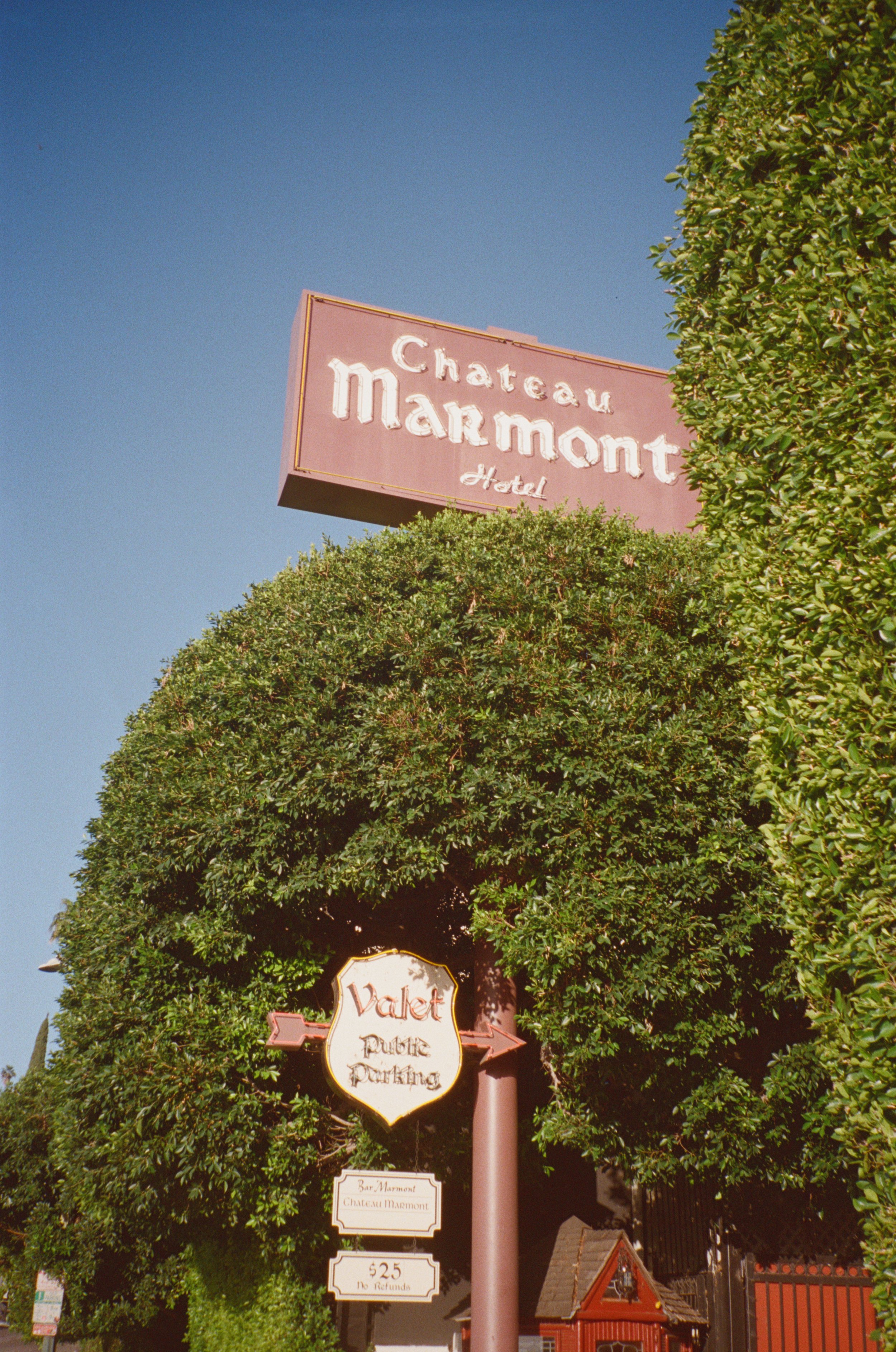 Sign for Château Marmont Hotel and Valet Parking amid lush green trees, with clear blue sky in the background.