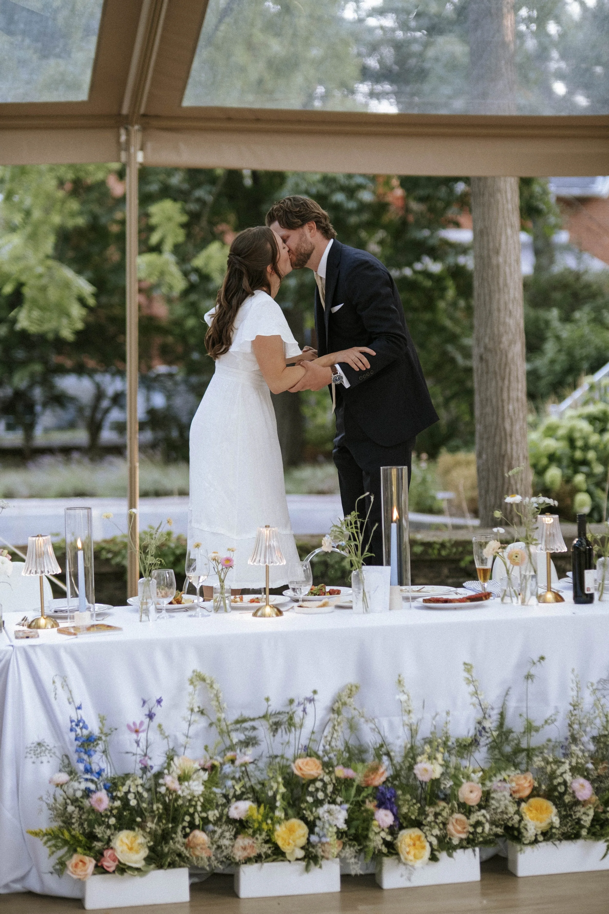A bride and groom sharing a kiss during their wedding reception under a canopy, with a decorated table and colorful flowers in the foreground.