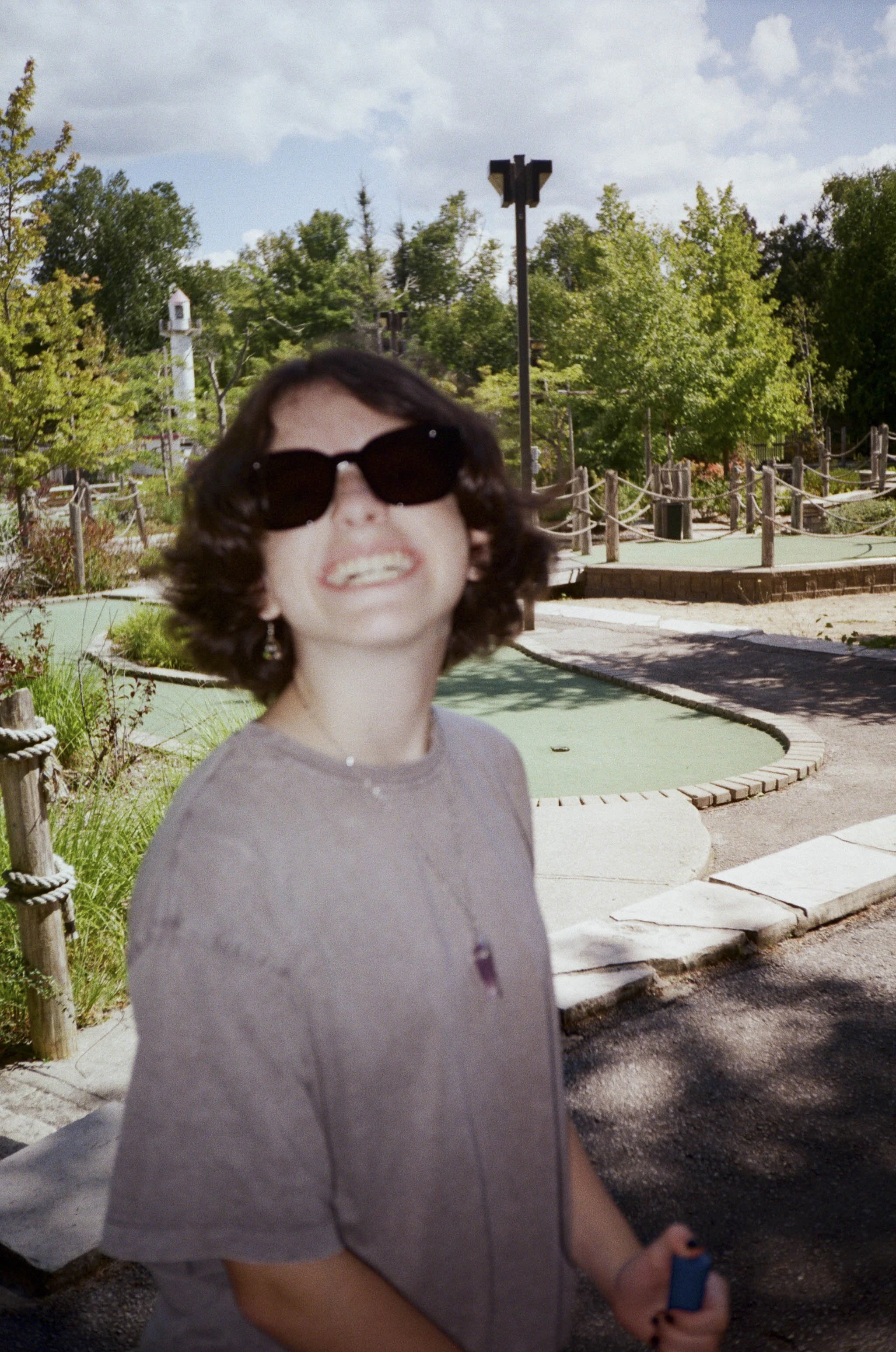 A woman wearing large black sunglasses and a light-colored shirt, smiling outdoors on a sunny day, with greenery and a mini-golf course in the background.
