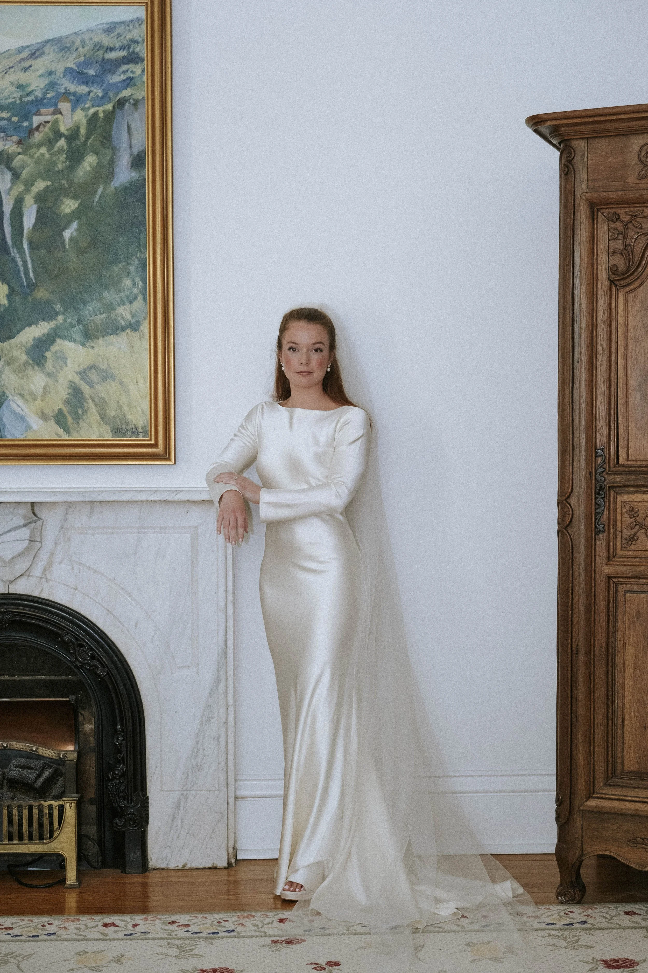 A woman in a long, elegant, white satin gown standing beside a marble fireplace and wooden cabinet in a room with a painting on the wall.