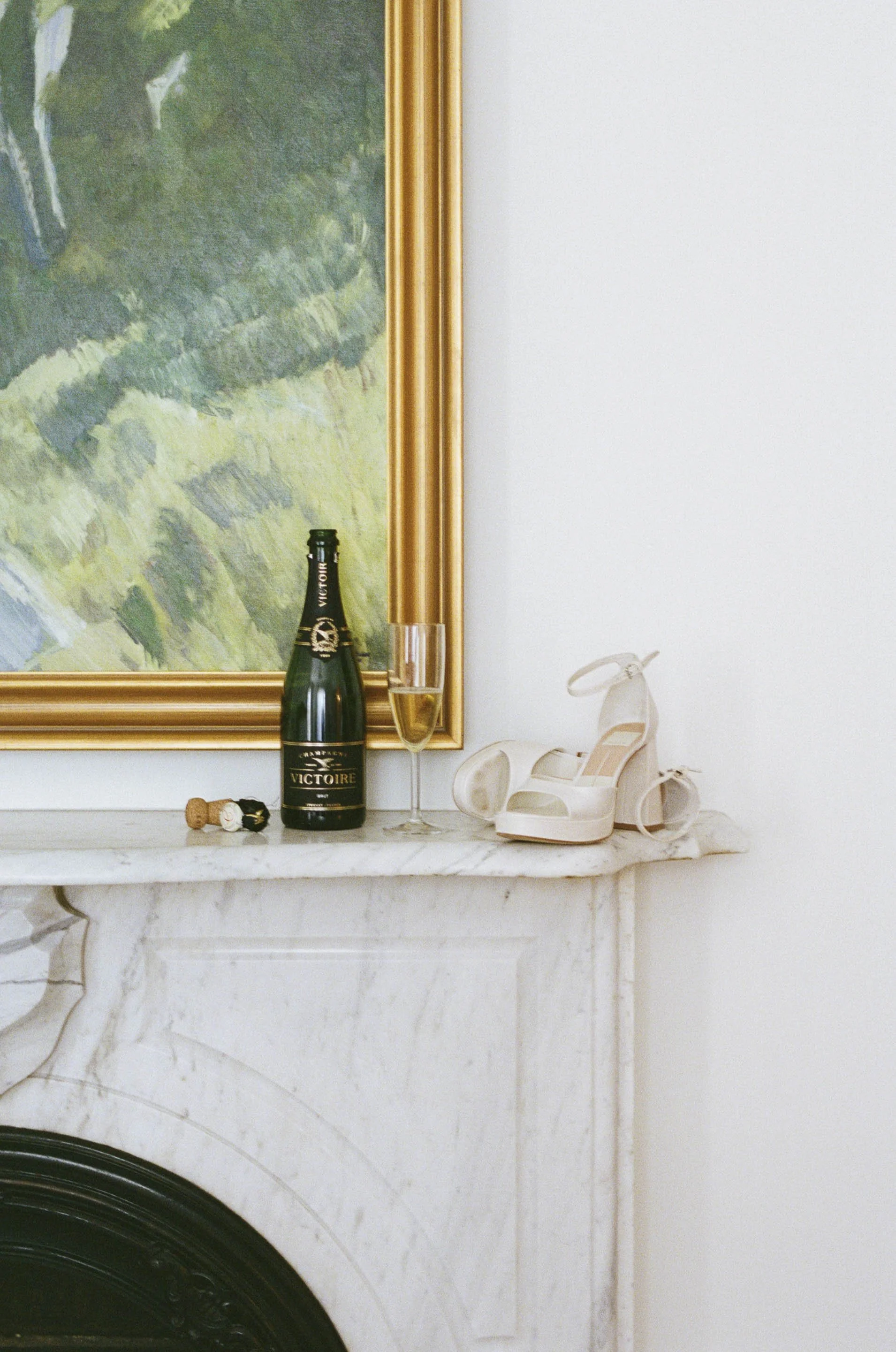 A champagne bottle, a glass of champagne, a pair of white high-heeled shoes, a cork, and a small accessory on a fireplace mantel with a large framed painting in the background.
