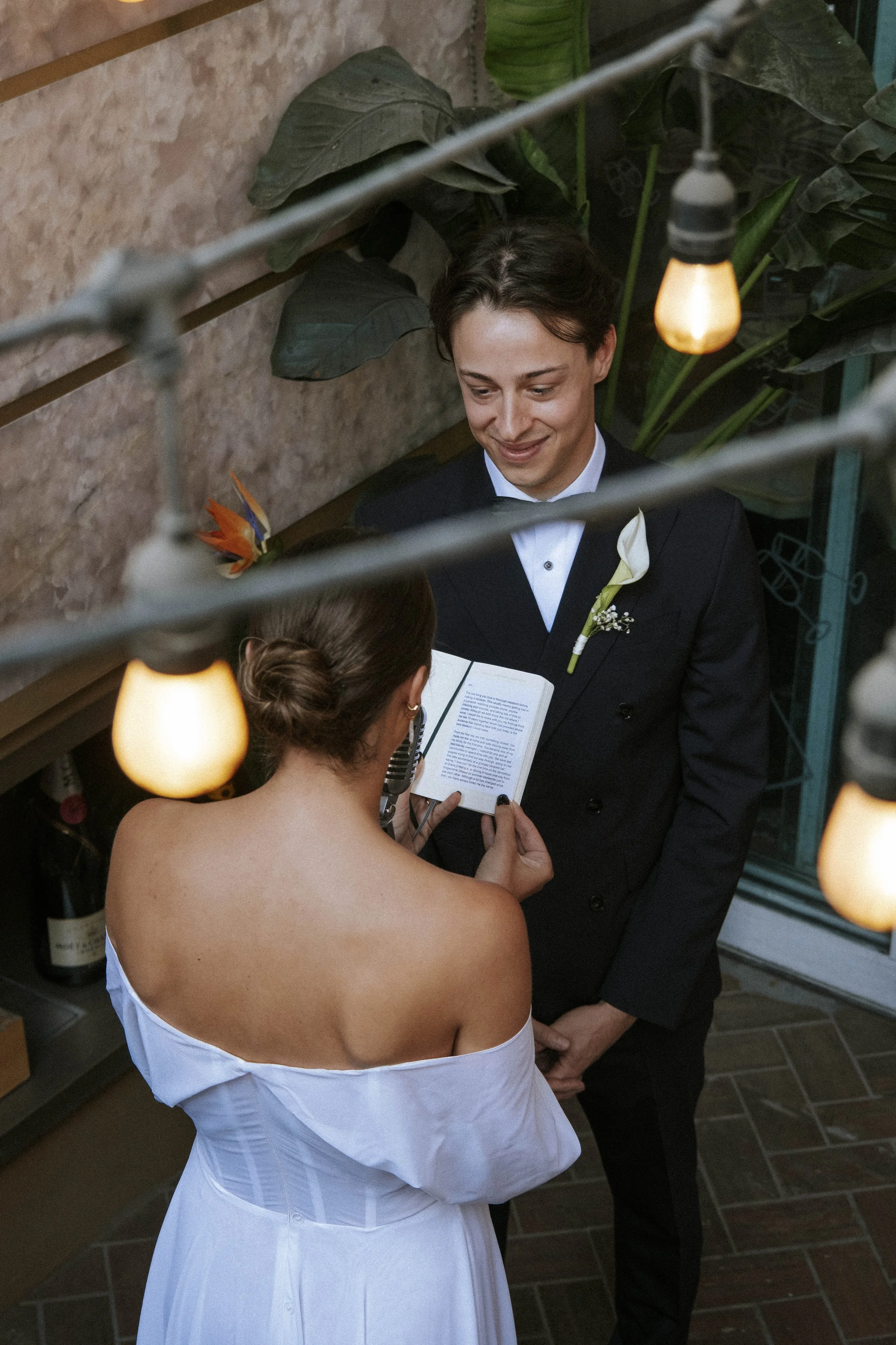A bride and groom exchanging vows during a wedding ceremony, with the bride reading from a small book and the groom smiling. The setting is decorated with string lights and green plants.
