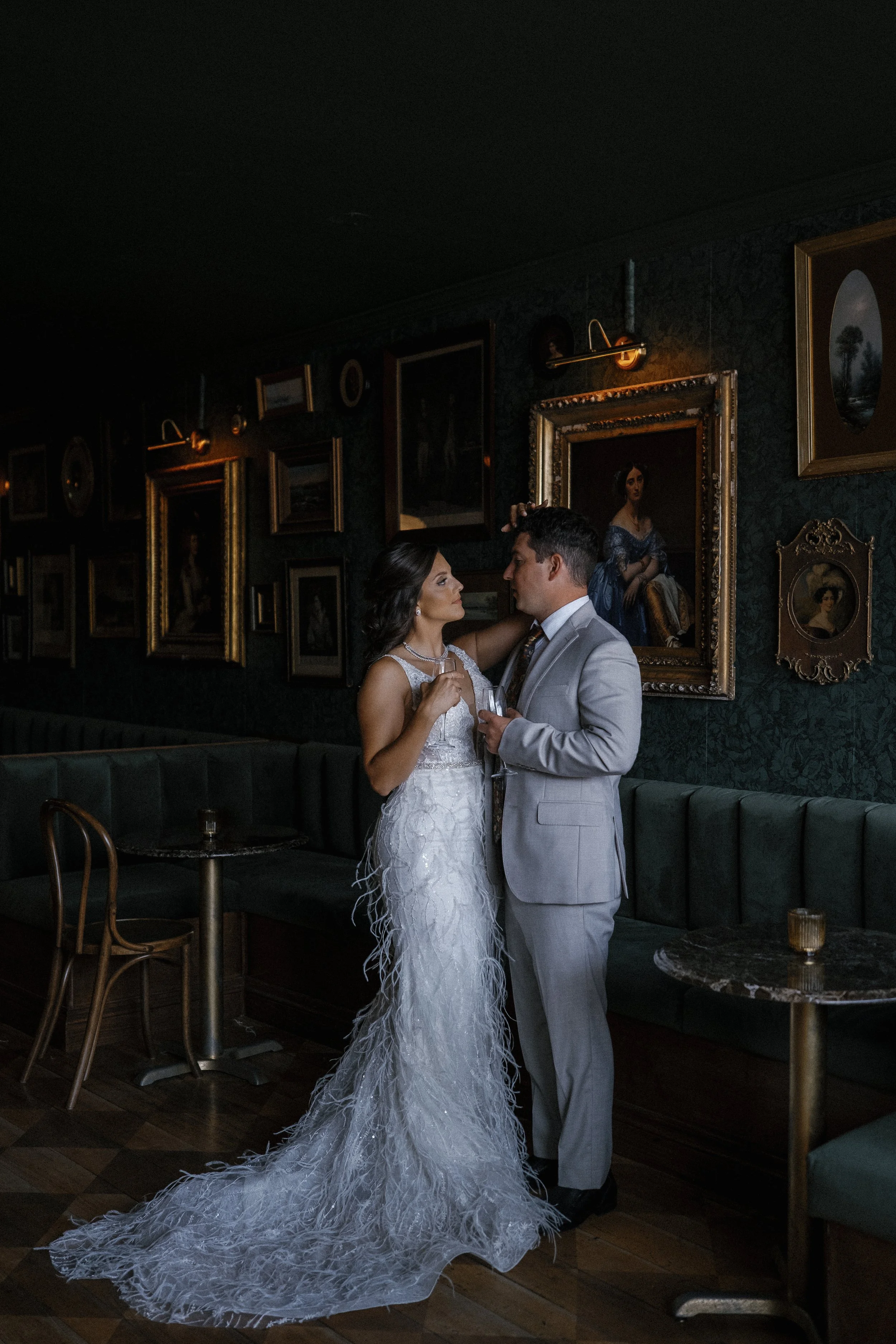A bride and groom stand close together inside a dimly lit, art-filled room, gazing into each other's eyes. The bride is wearing an elegant white gown with feathered details and holds a glass. The groom is in a light gray suit with no hat, holding a g