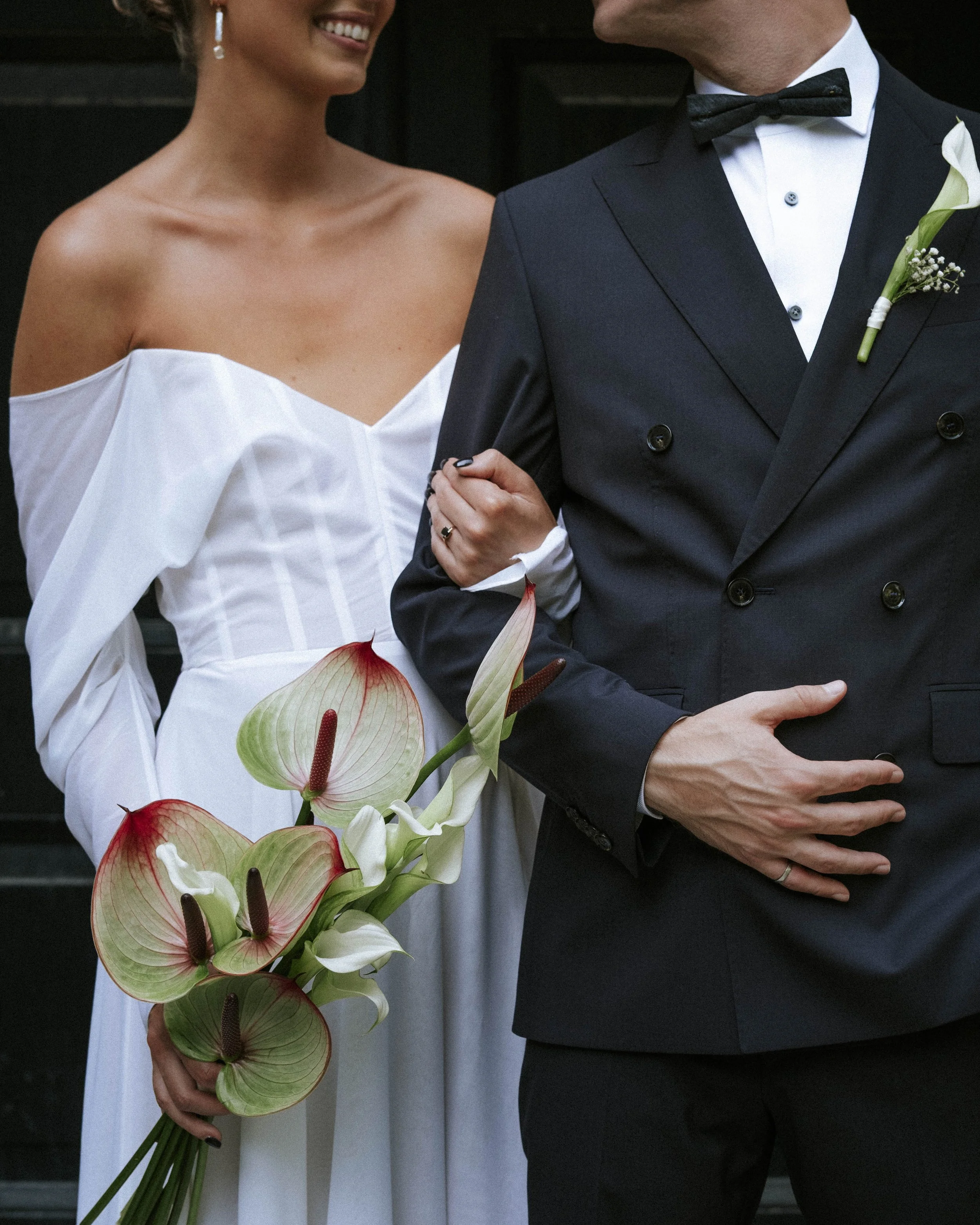 A bride and groom on their wedding day, with the bride holding a bouquet of white and red calla lilies, wearing a white off-the-shoulder wedding gown, and the groom dressed in a dark tuxedo with a bow tie and boutonniere.