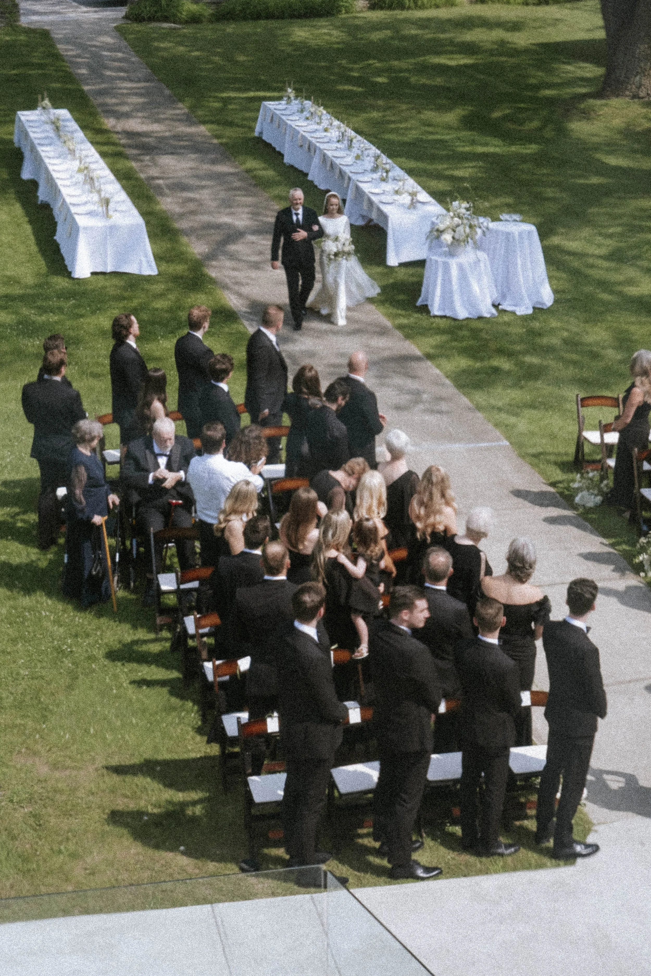 People attending a wedding ceremony outdoors, with the bride walking down the aisle escorted by a man, likely her father, toward the officiant. Guests are seated in rows on either side of the aisle, with some standing and watching the procession.