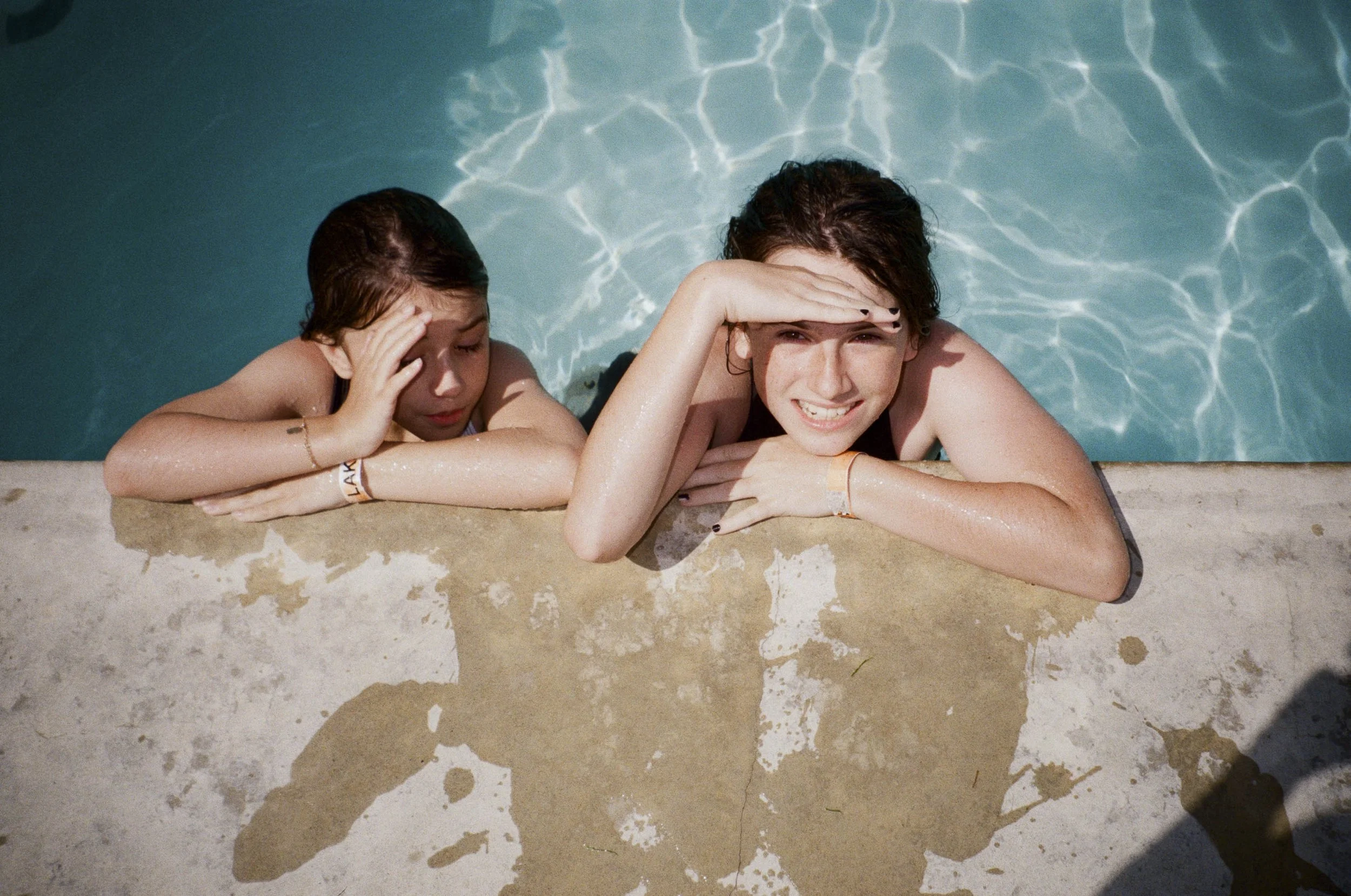 Two young women resting on the edge of a swimming pool, sunbathing and shielding their eyes from the sun with their hands.