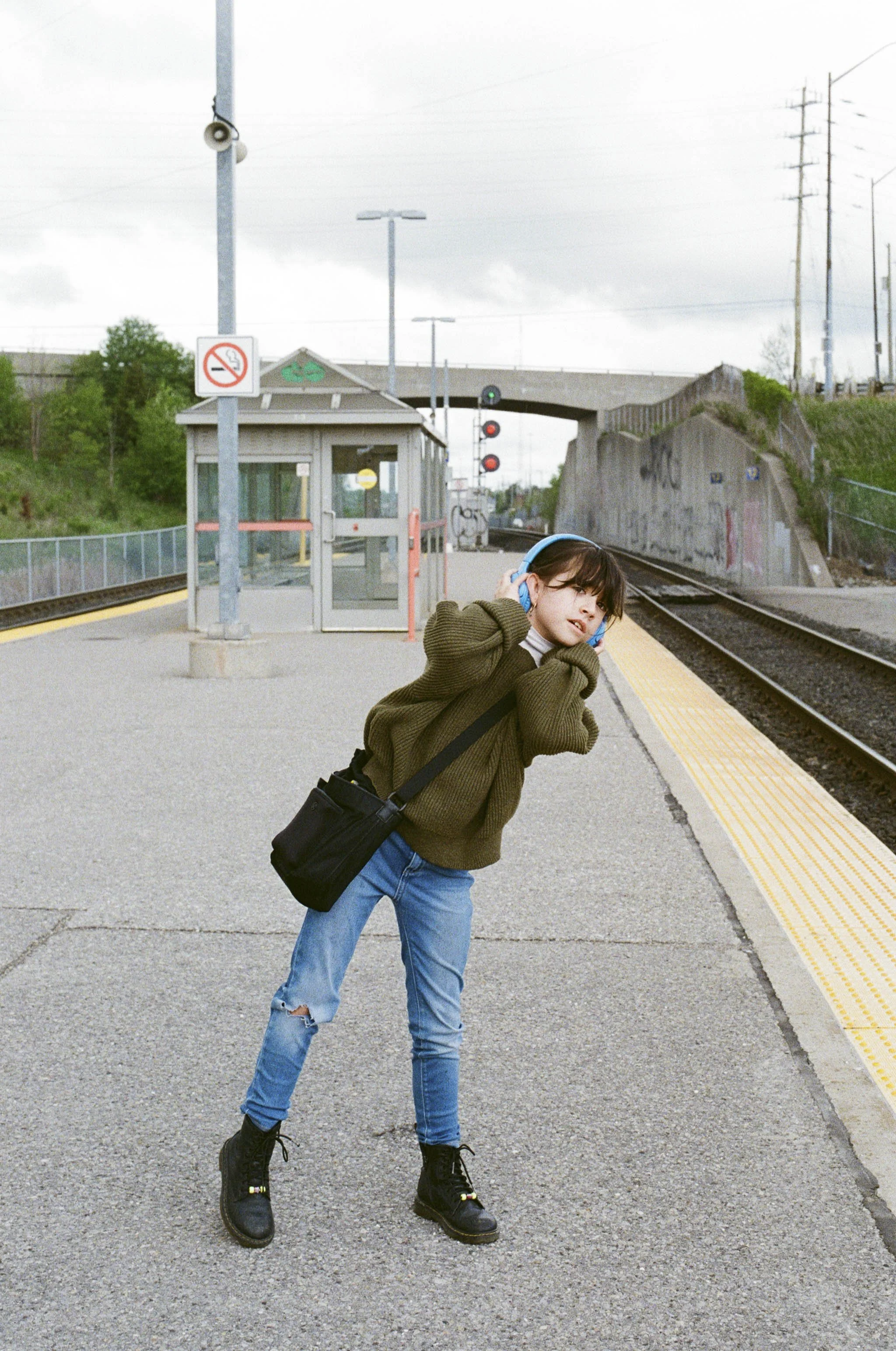 A young person at a train station wearing headphones, a brown sweater, ripped jeans, and black boots, holding their ears and leaning back.