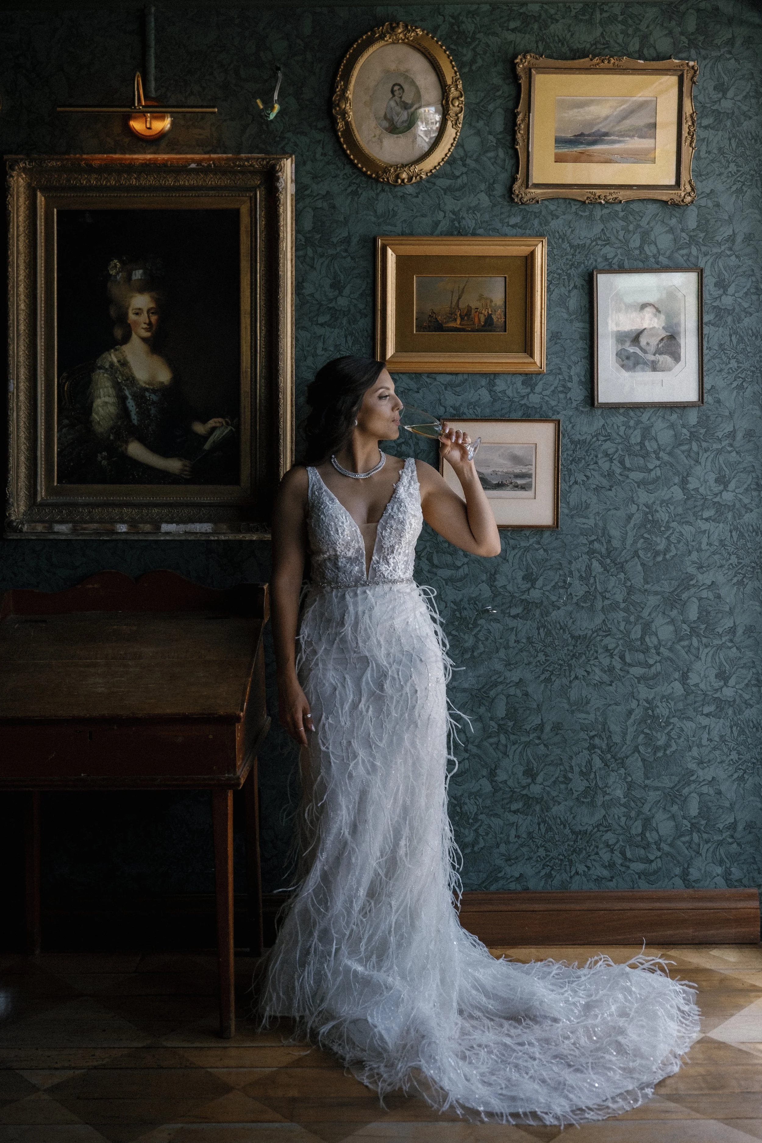 A woman in a white wedding dress is standing in a room with green floral wallpaper, drinking from a glass, and surrounded by framed paintings and portraits.