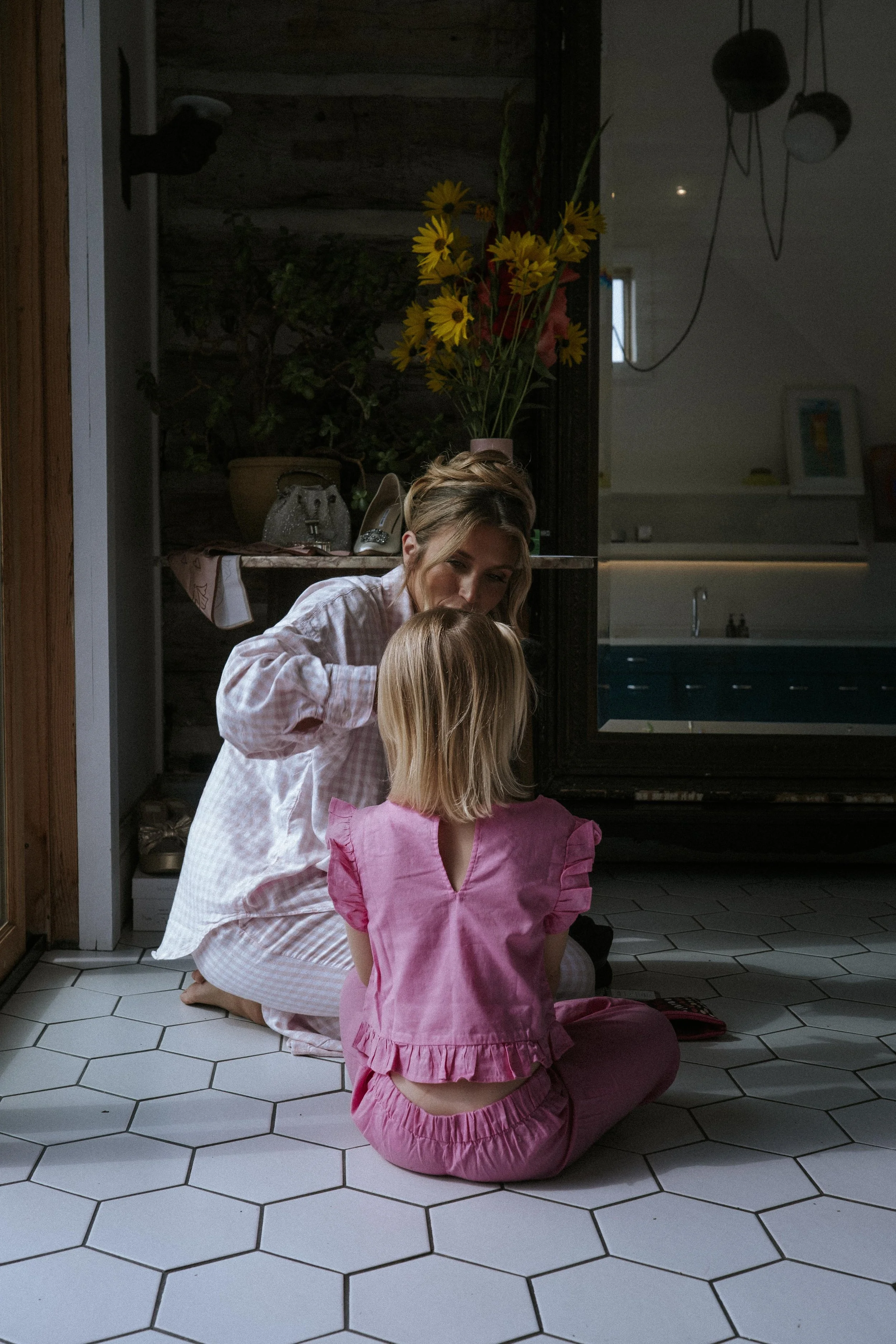 A woman and a young girl sitting on a tiled floor inside a house, facing each other. The woman is leaning forward, talking to the girl, who is wearing pink pajamas. There is a large mirror behind them and a flower arrangement in a pink vase on top of a shelf near the mirror.