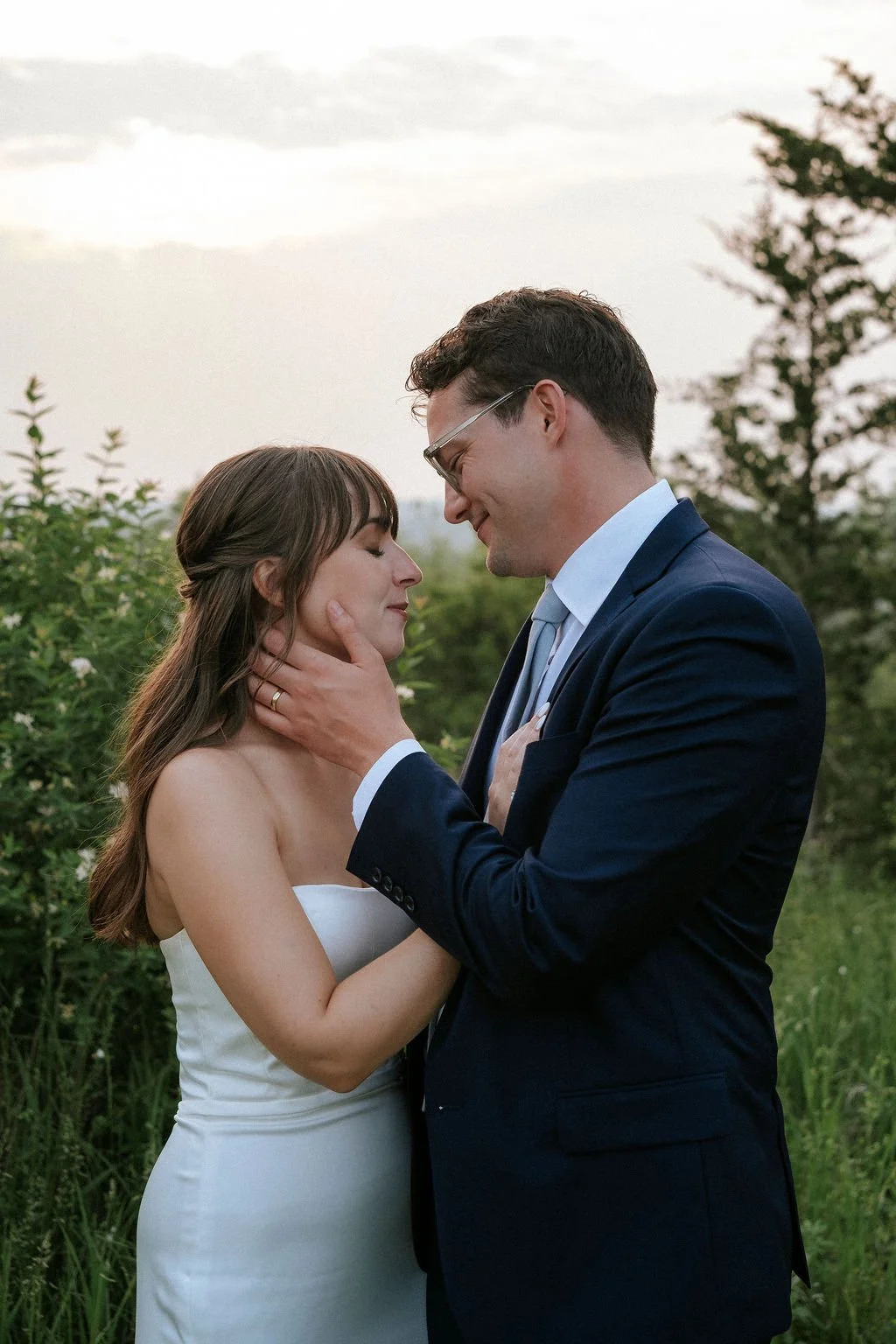 A bride and groom sharing an intimate moment outdoors, with the groom gently holding the bride's face and the bride smiling softly with her eyes closed.