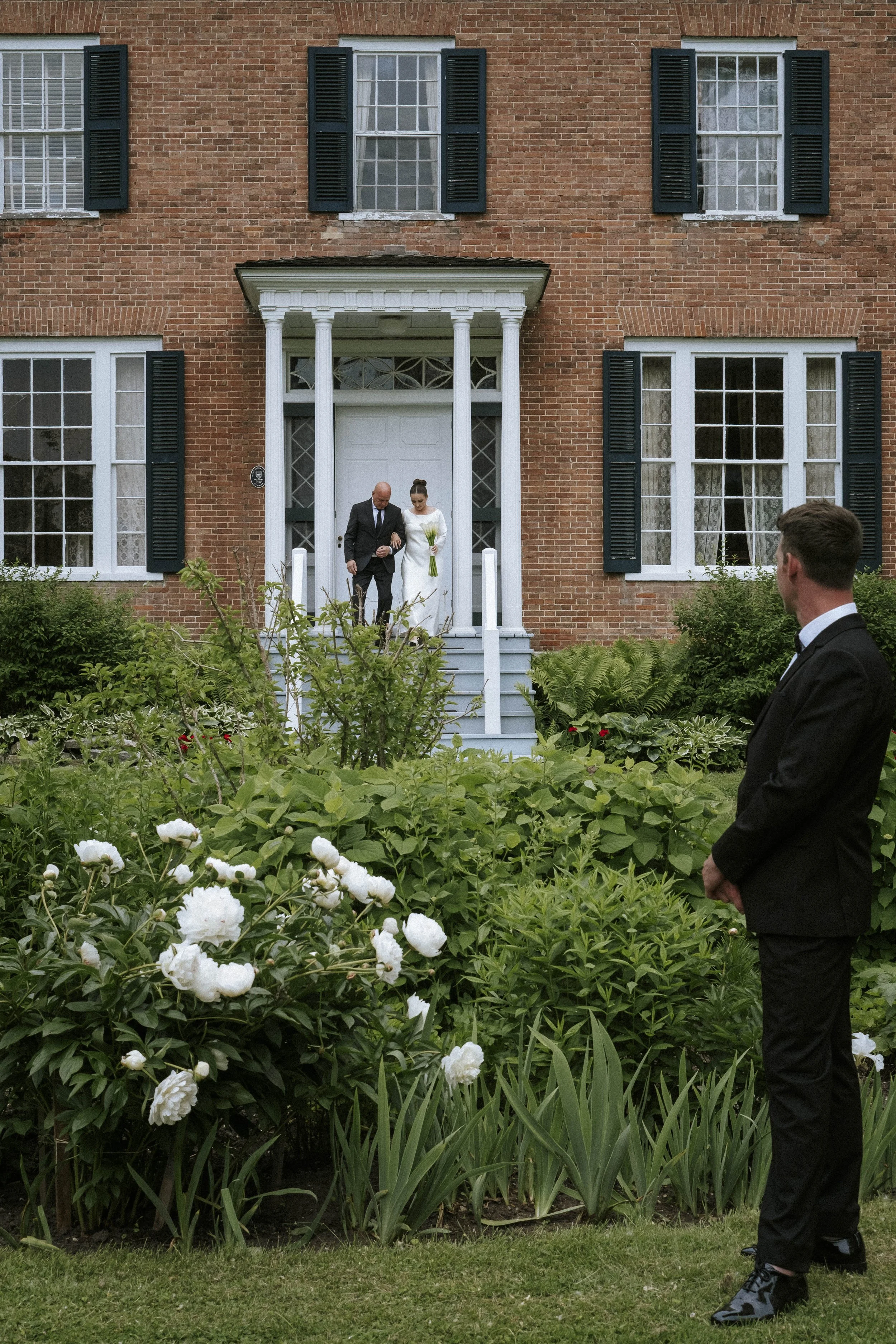 A bride walking down the steps of a brick house with a man, possibly her father, while a man in a black tuxedo watches from the garden with white flowers.