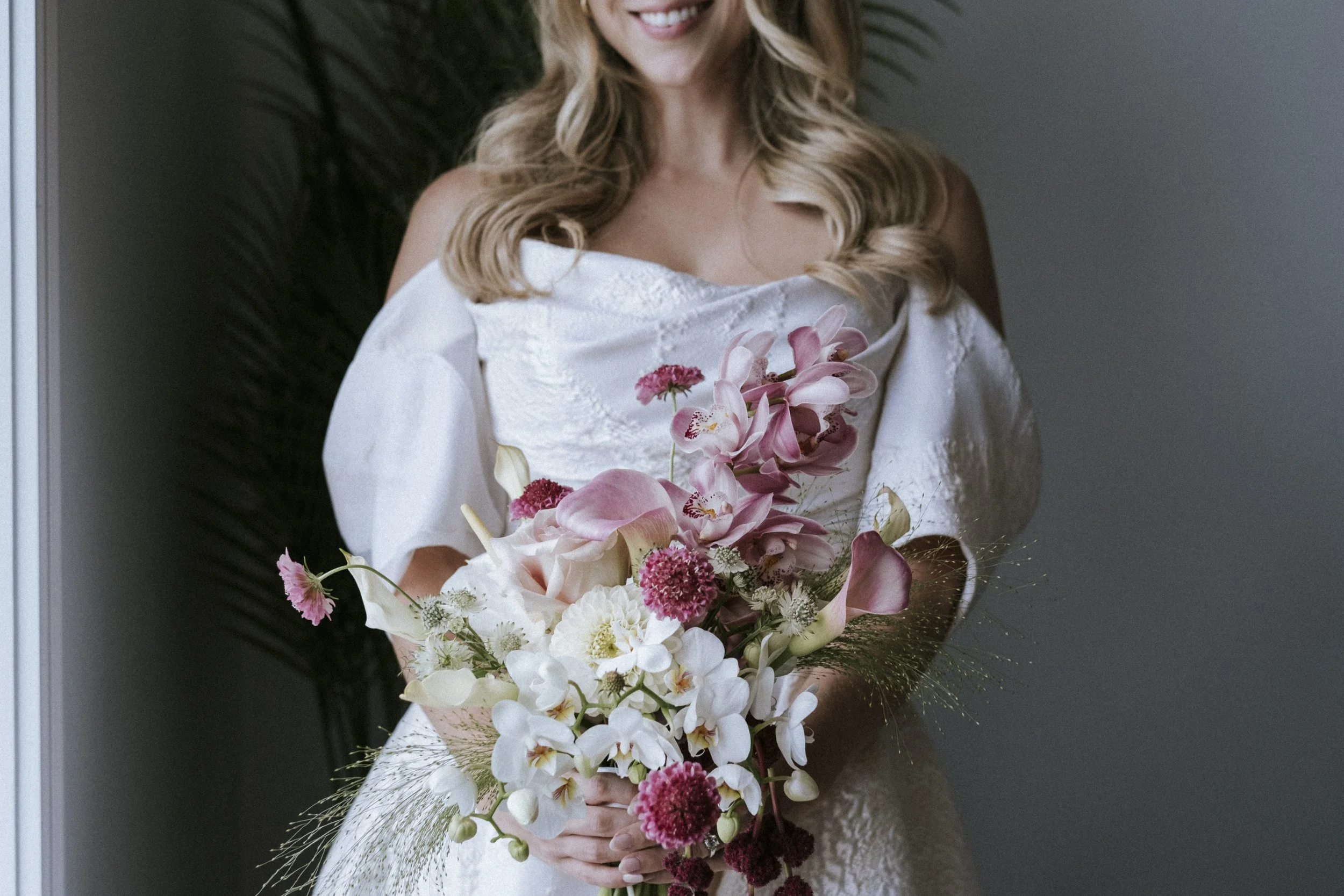A woman in a white dress holding a large bouquet of pink and white flowers, with a smiling man standing behind her