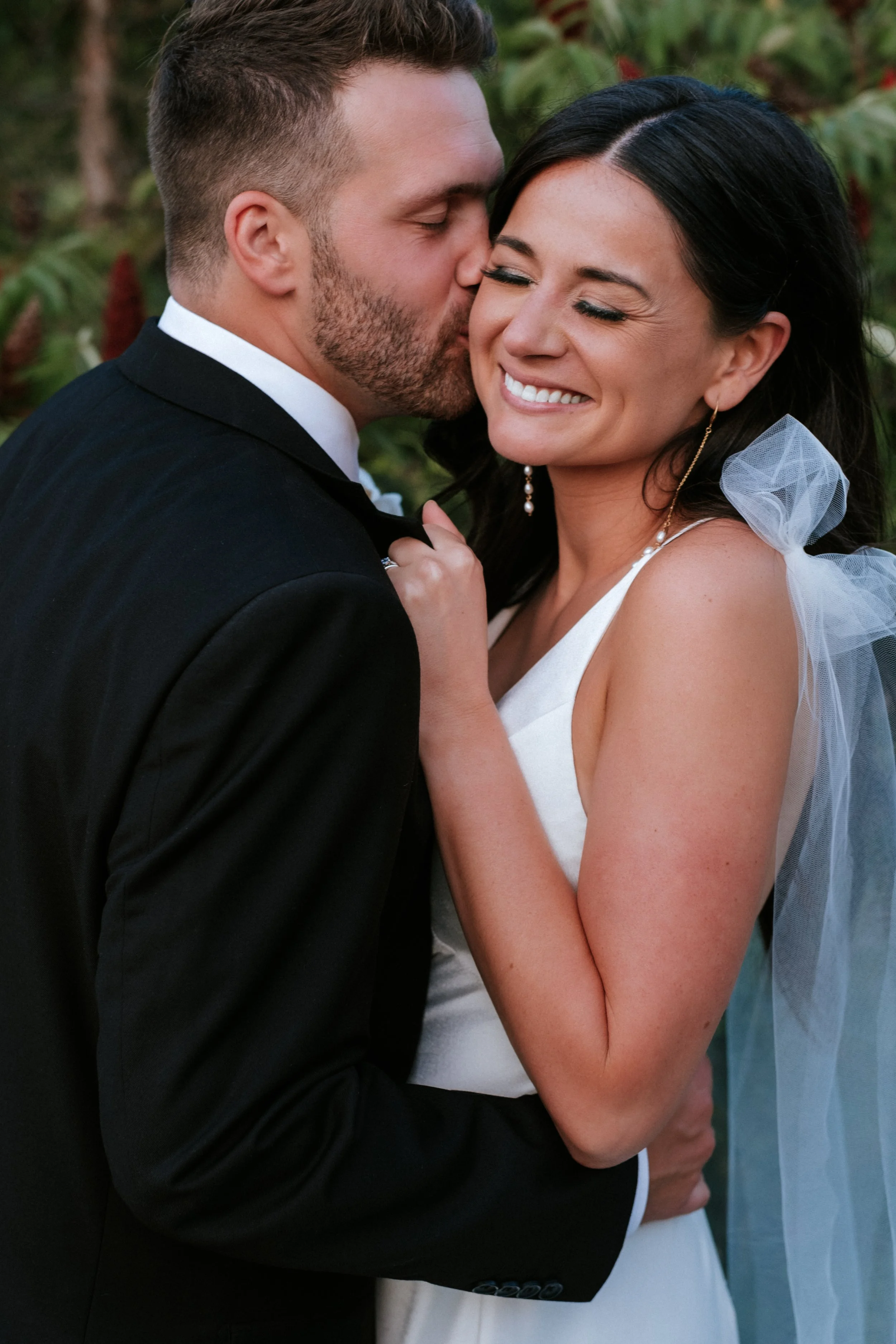 A newlywed couple embraces, with the man kissing the woman on her cheek and the woman smiling with eyes closed. The woman is wearing a white wedding dress and veil, and the man is in a black tuxedo. They are outdoors with green foliage in the backgro
