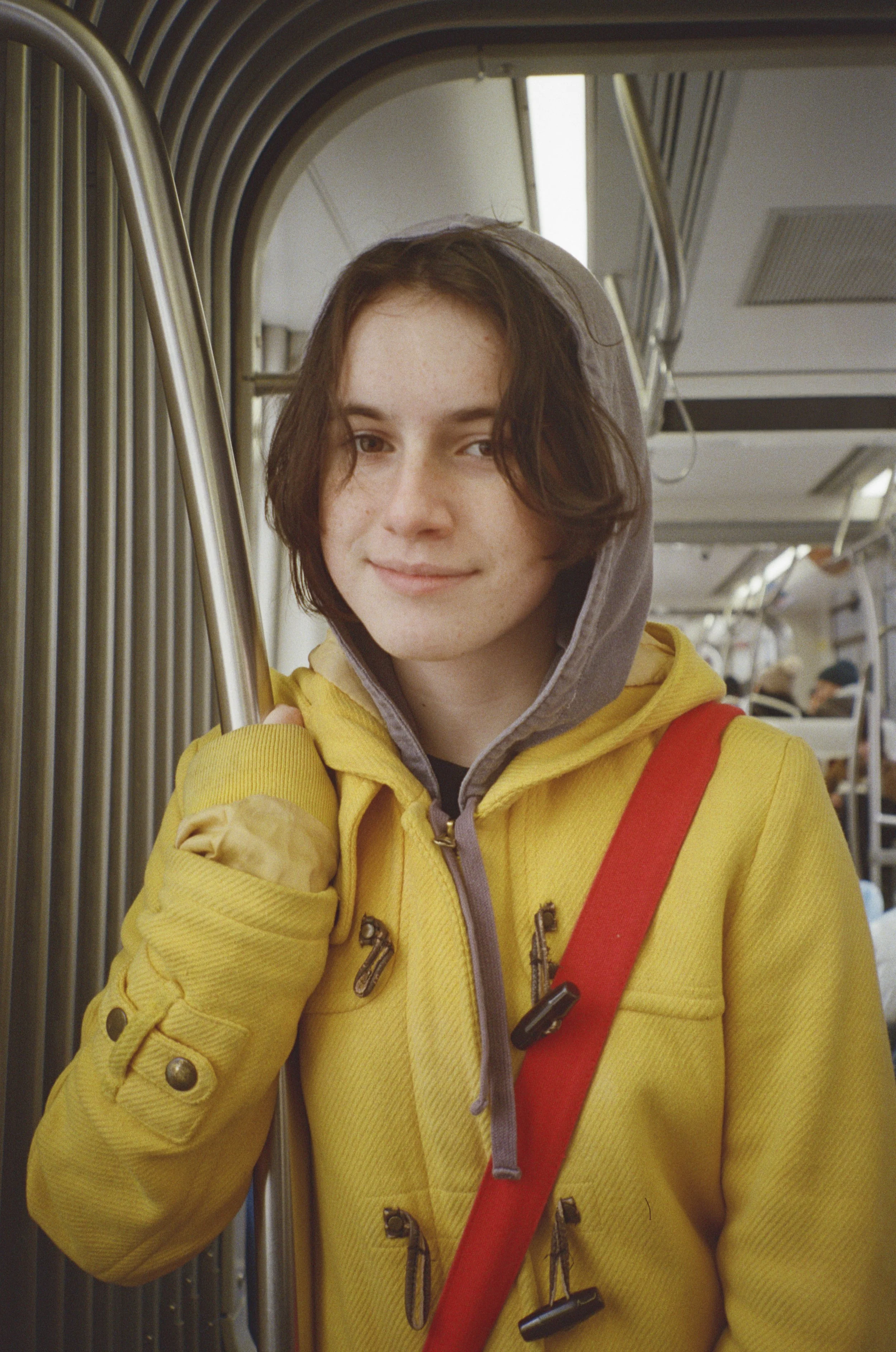 A young woman with short brown hair and a slight smile, wearing a yellow hoodie with a purple lining, holding onto a metro or subway handrail inside a train.
