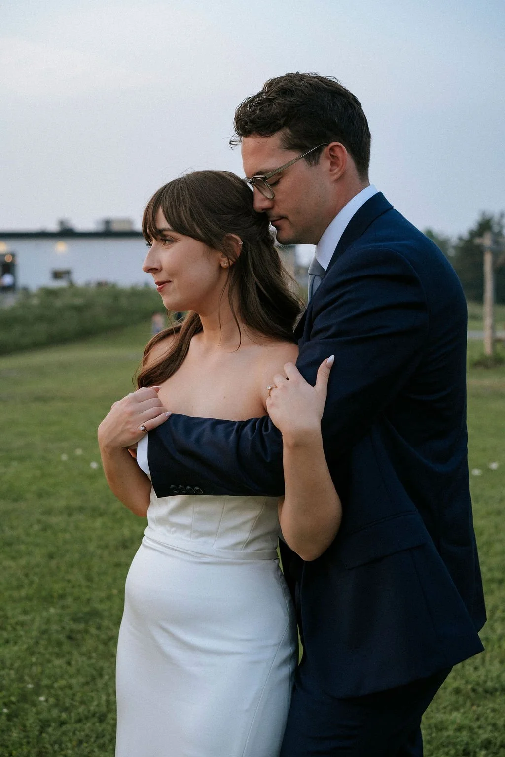 A couple in formal attire embracing outdoors on a grassy field, with a building and trees in the background during evening time.