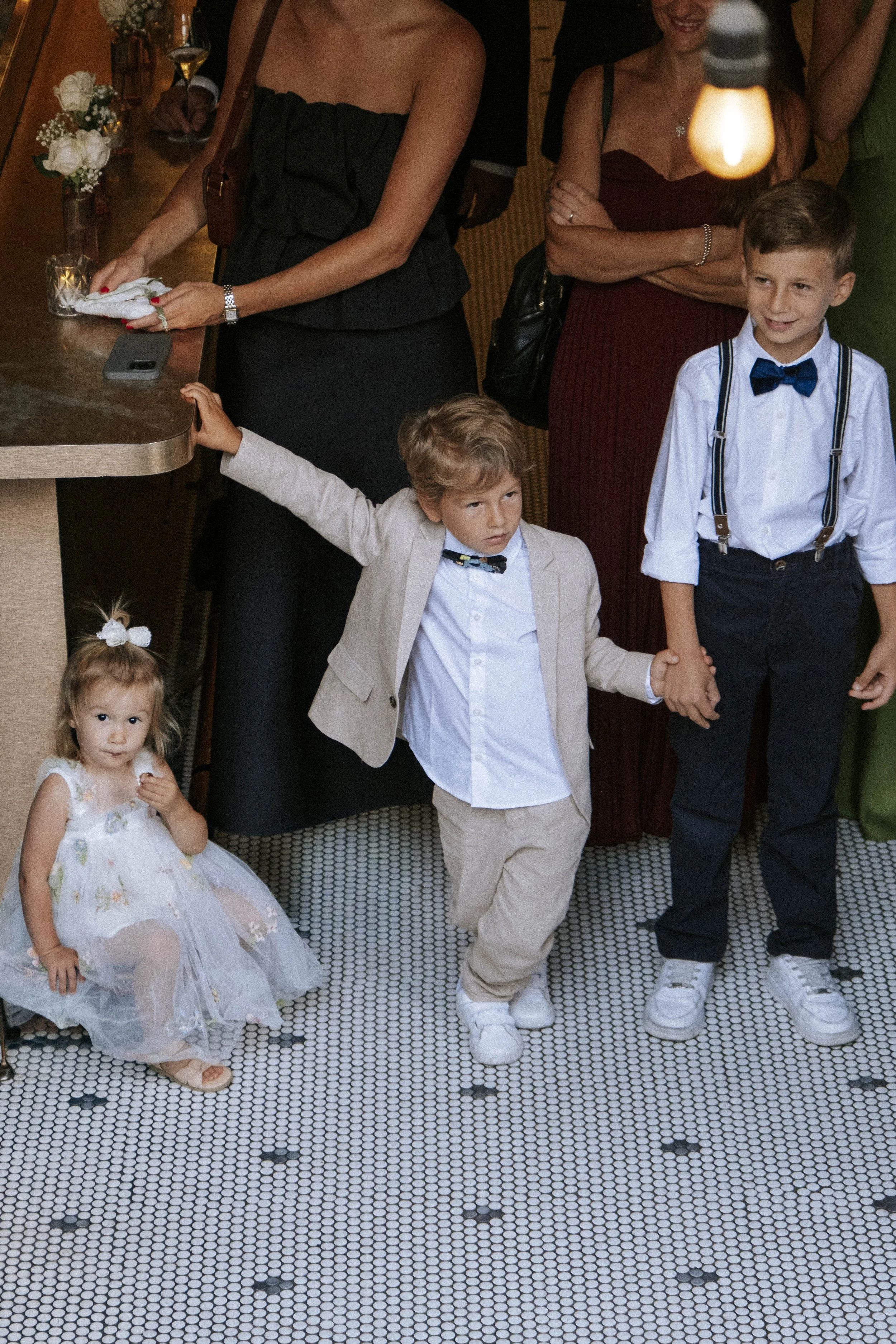Three children, two boys and a girl, are dressed formally at a social event. The girl is sitting on the floor in a floral dress. The boys are holding hands, with one wearing a beige suit and the other wearing a white shirt with suspenders and a bow tie. Adults are standing nearby, some with drinks, in a warmly lit room with a tiled floor and some floral decorations on a counter.