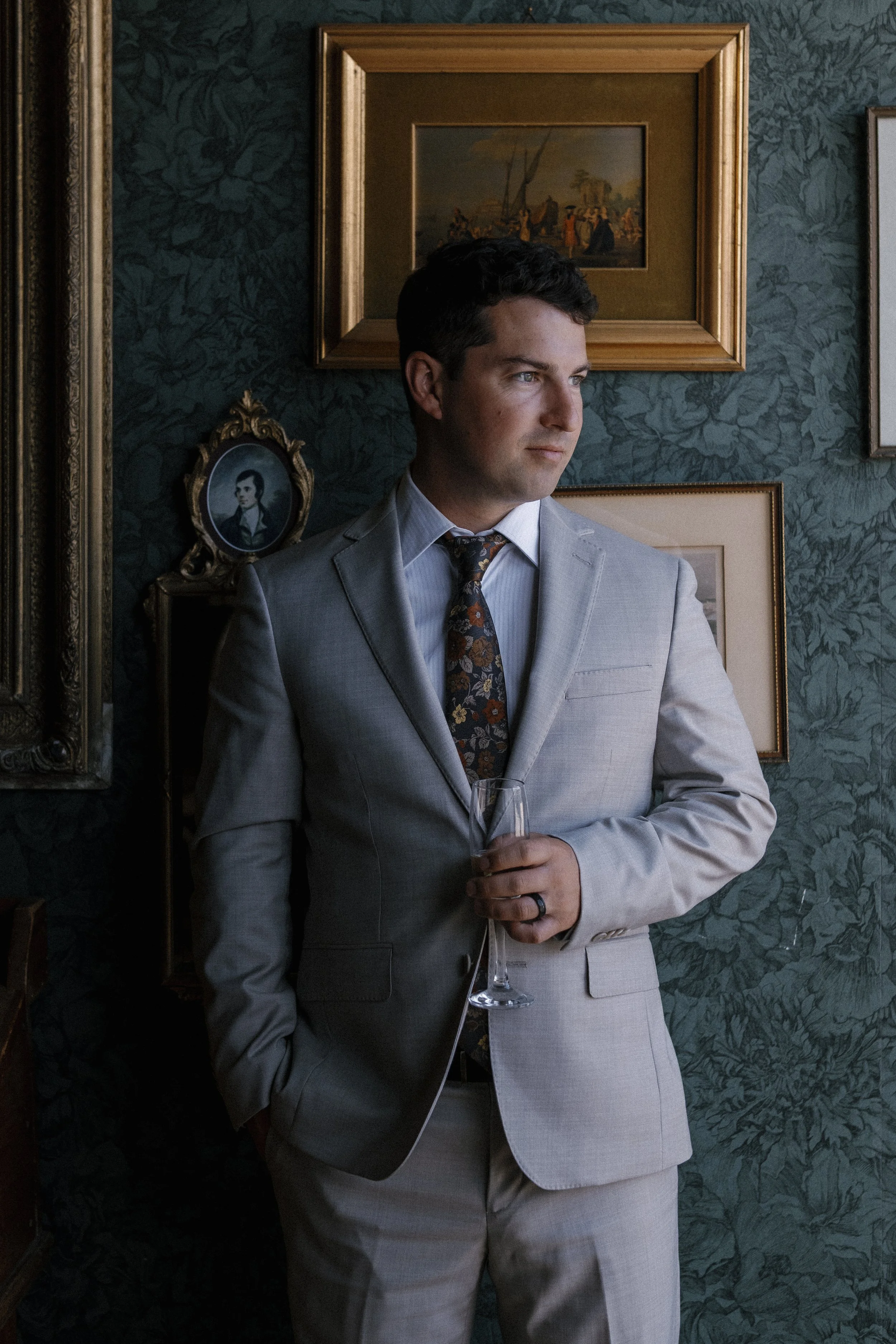 A man in a light gray suit holding a glass, standing against a dark floral wallpapered wall decorated with framed artwork and portrait photographs, looking to the side.