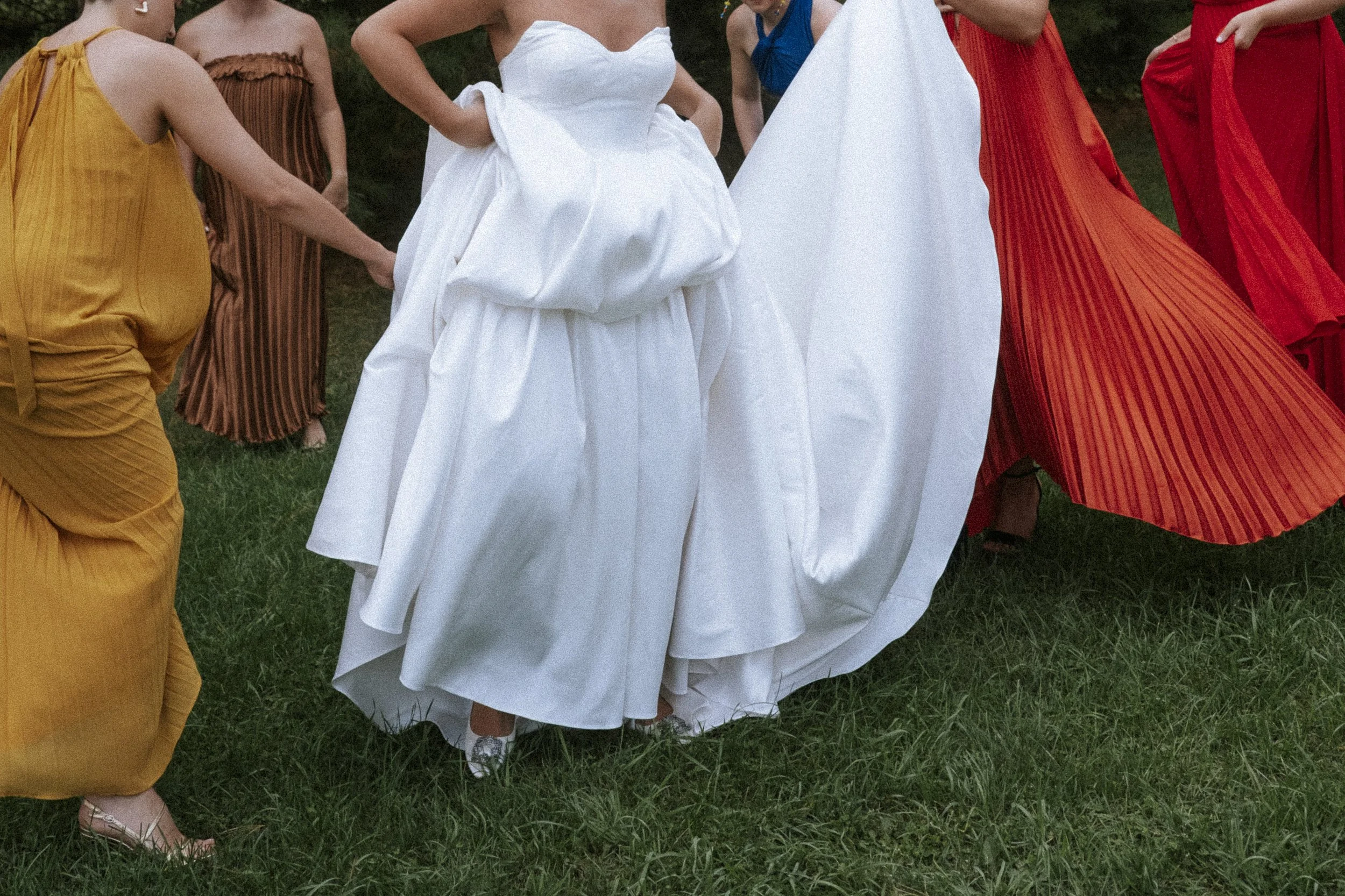 A group of women wearing colorful dresses, with one woman in a white dress, standing on grass in an outdoor setting.