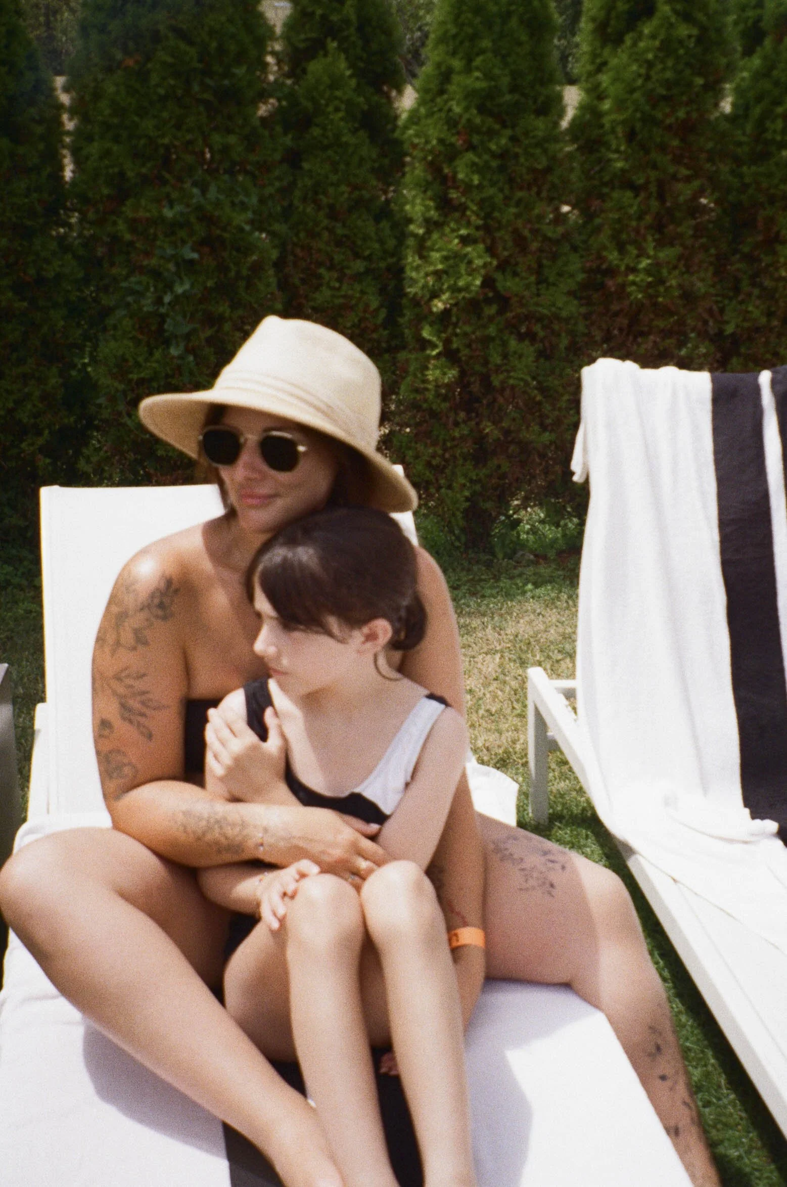 Woman and girl sitting on a lounge chair outdoors, woman wearing a large sun hat and sunglasses, girl with short dark hair, woman hugging girl, towel draped over another lounge chair nearby, green bushes in the background.