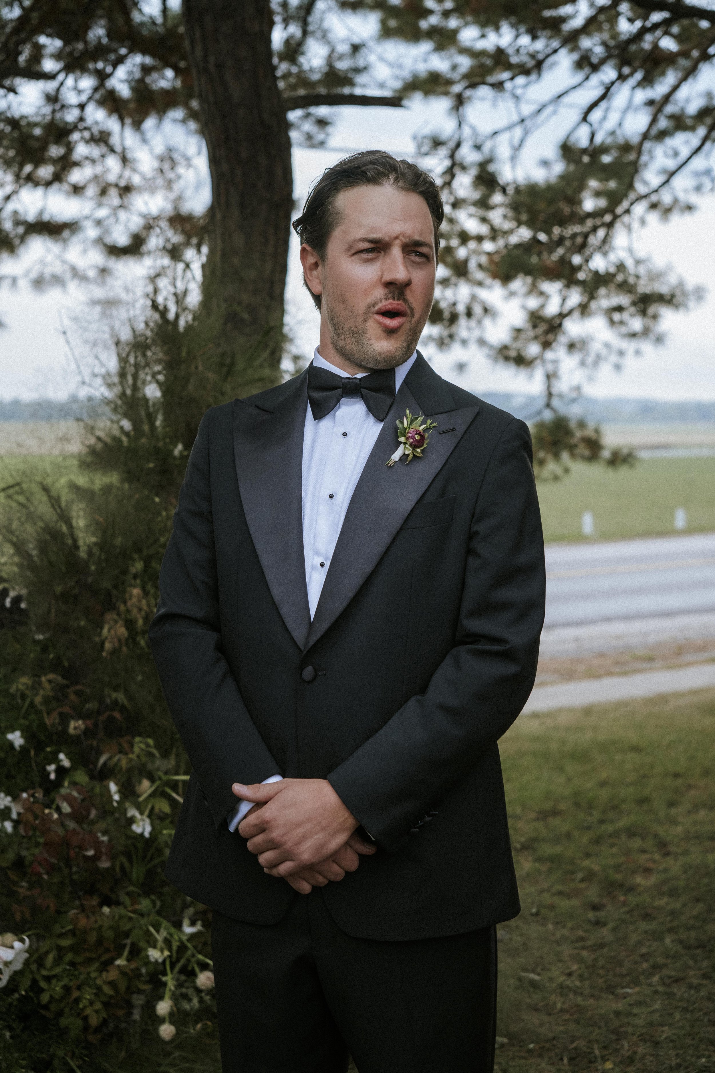 A man in a black tuxedo with a bow tie, standing outdoors near a tree, with a serious or surprised expression, during daytime.