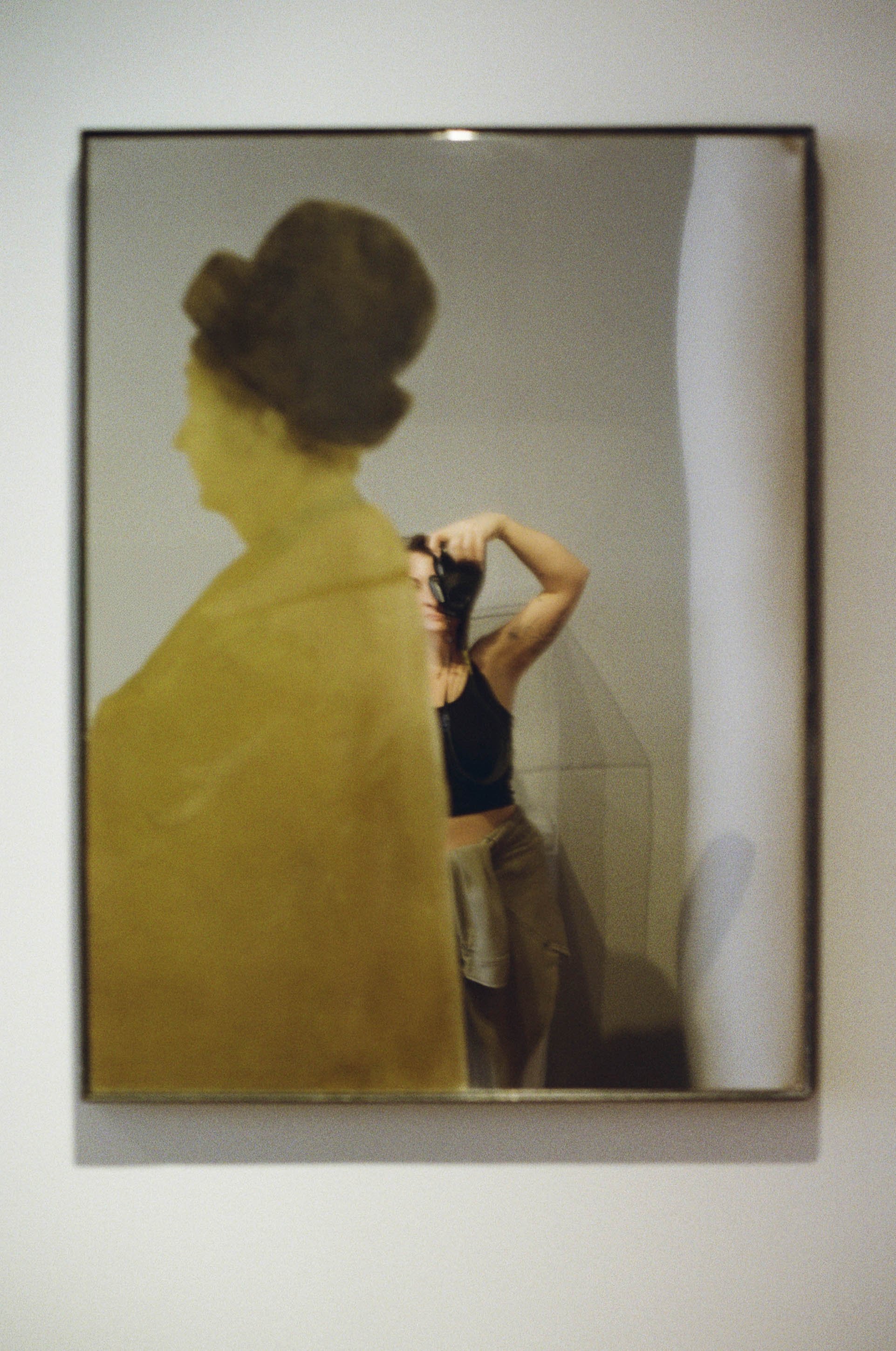 A woman taking a selfie in a mirror, with her arm raised and holding a camera, while her reflection is partially obscured by a yellow object.