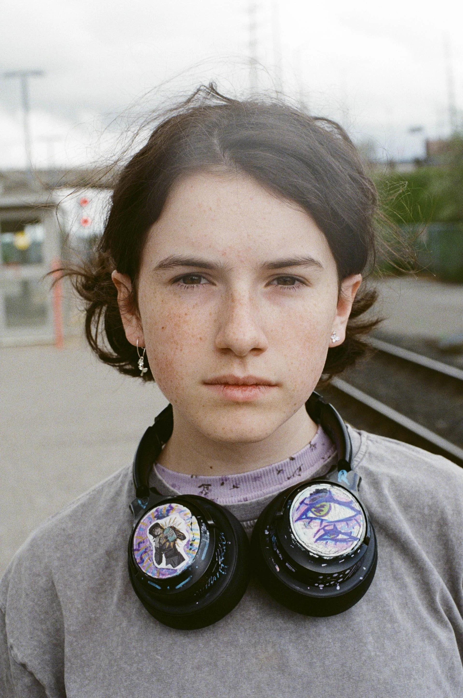 A young person with short dark hair, freckles, and earrings standing outdoors near train tracks, wearing headphones with colorful stickers.