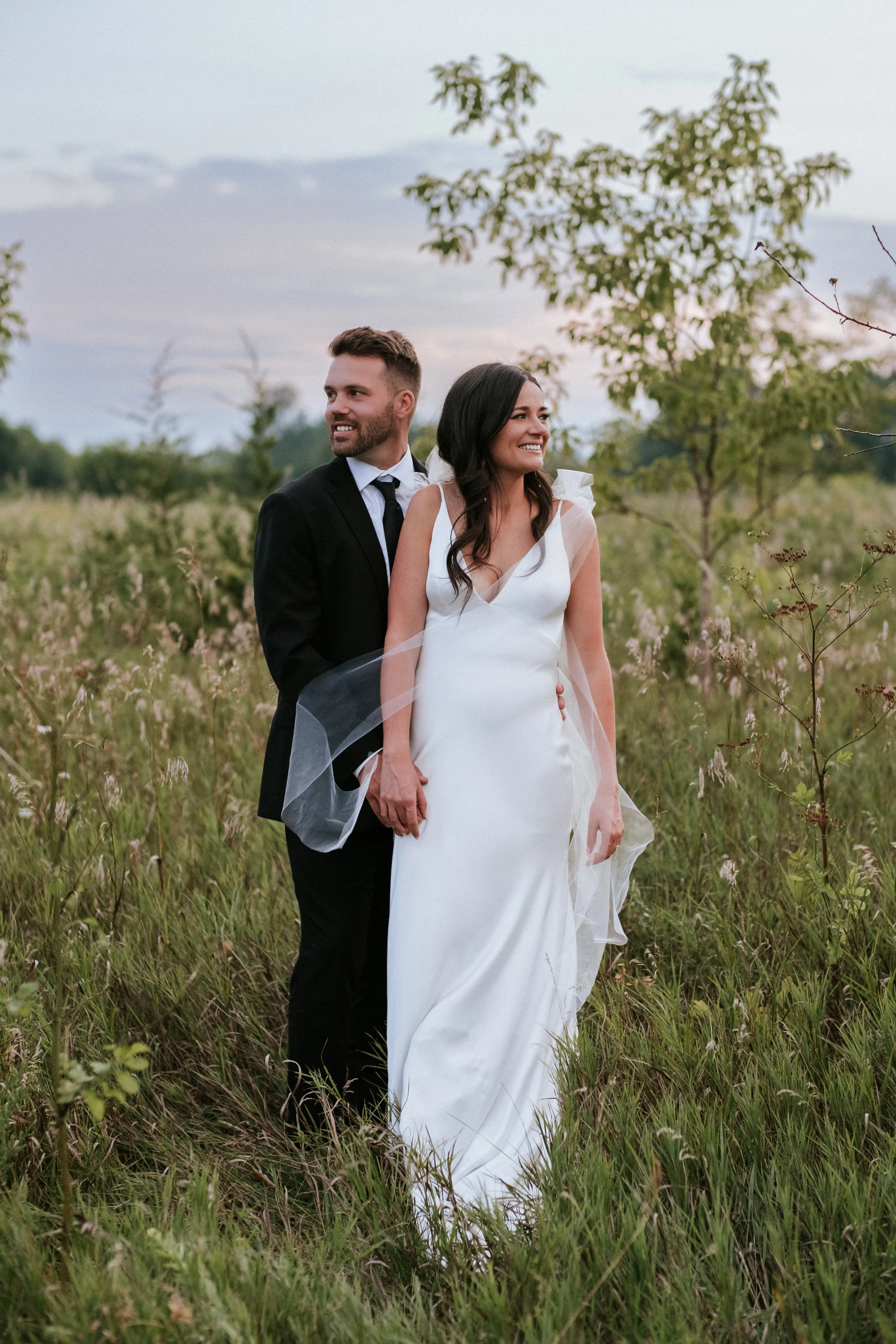 A bride and groom standing in a grassy field with trees in the background during sunset.