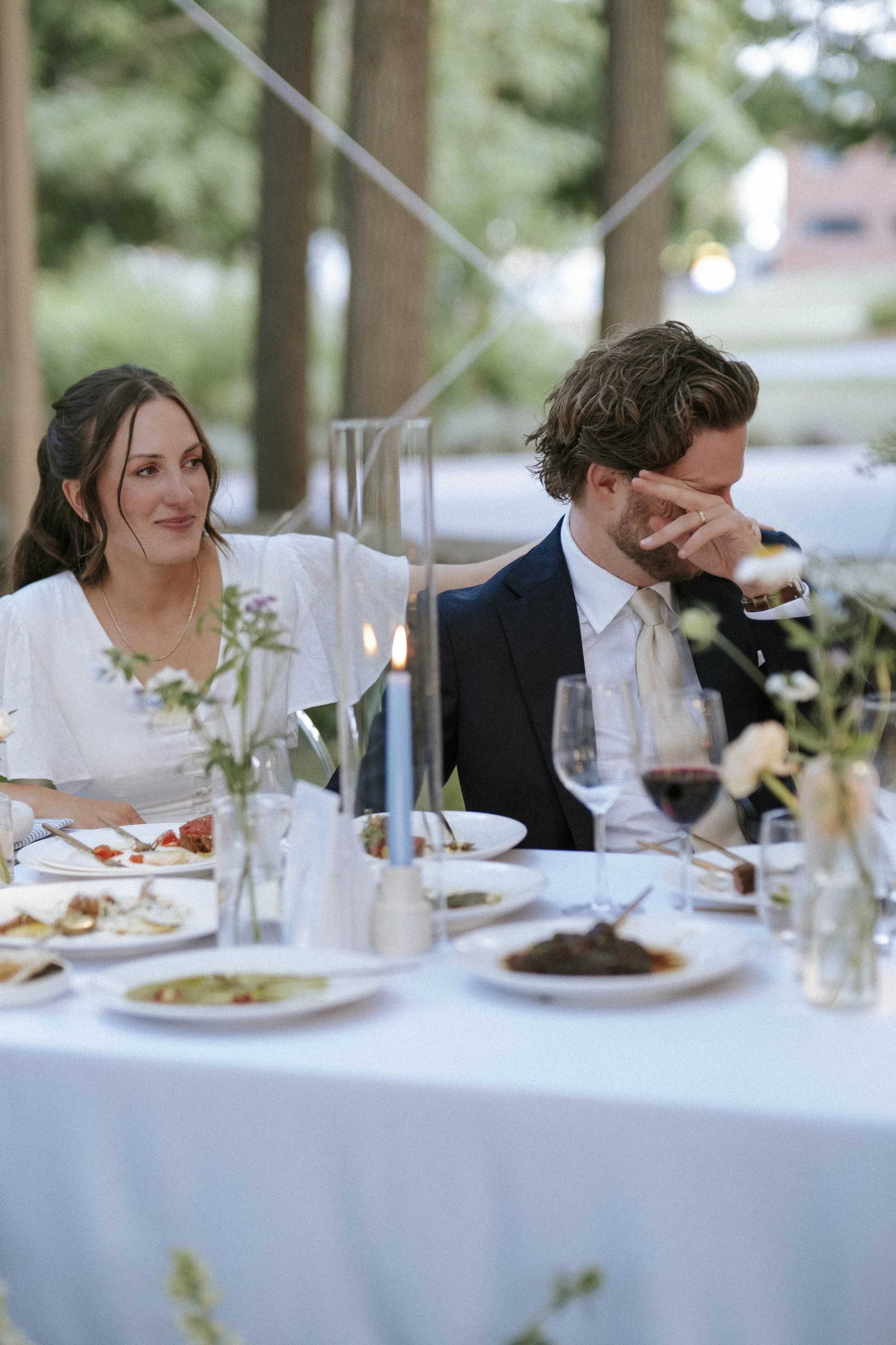 A woman in a white dress comforting a man with a beard in a tuxedo at an outdoor dinner table. The man appears emotional with his hand covering part of his face. The table is decorated with flowers, candles, and various dishes, with a blurred outdoor background of trees.