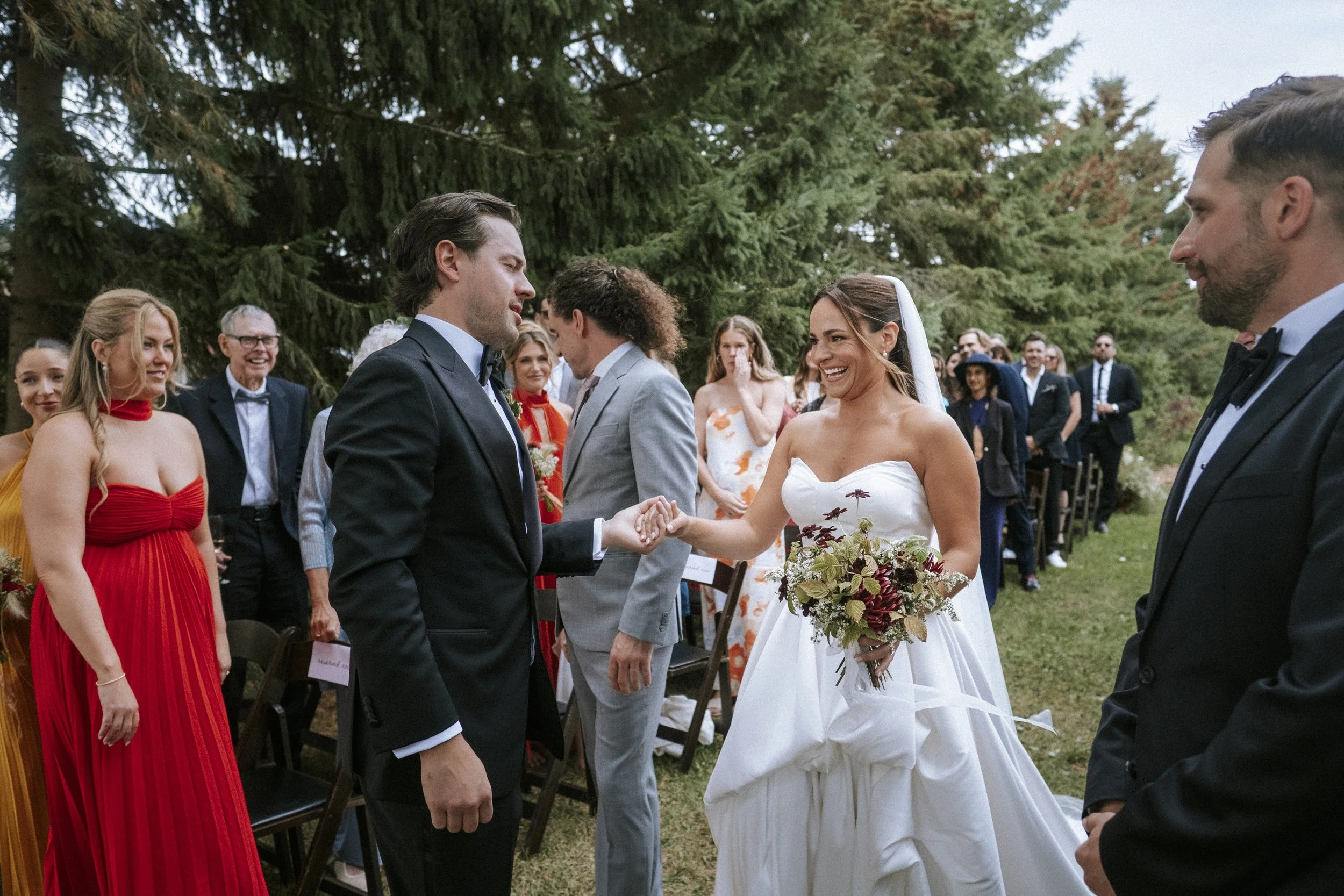 A bride and groom holding hands and smiling during their outdoor wedding ceremony surrounded by joyful guests, with trees in the background.