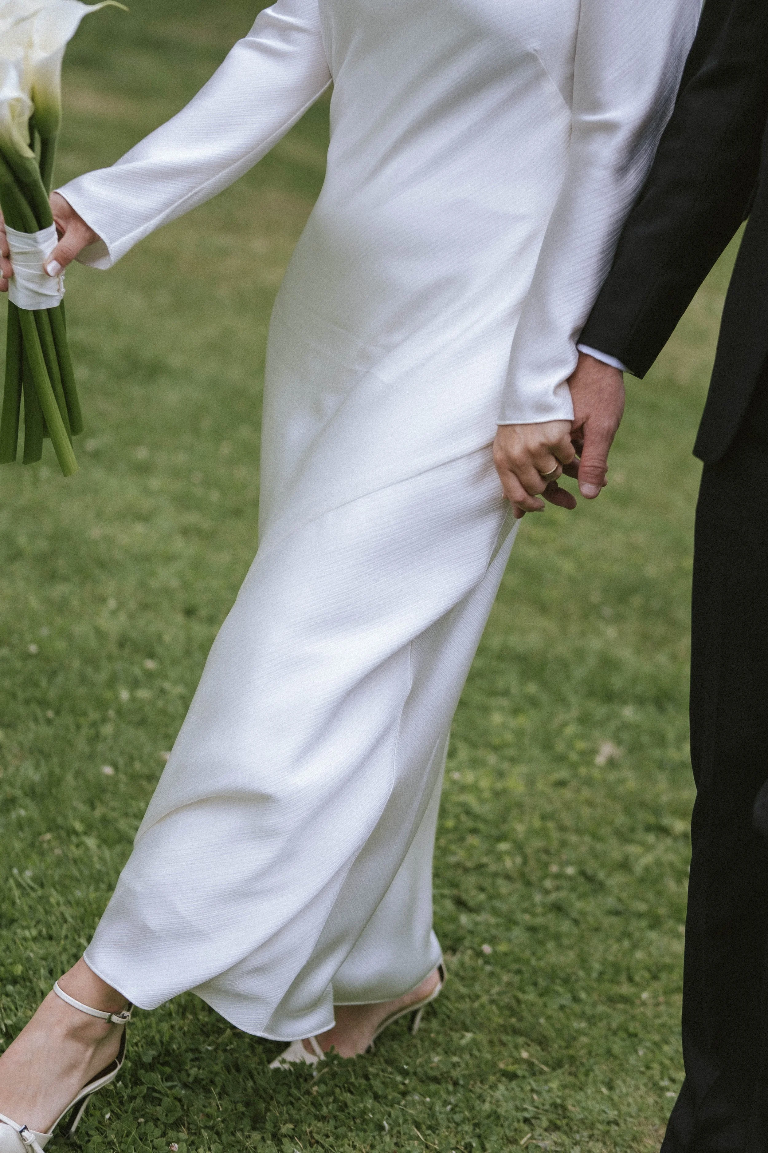 Close-up of a bride and groom holding hands during a wedding, bride wearing white high heels and a white dress, groom in black suit, on a grassy outdoor setting.