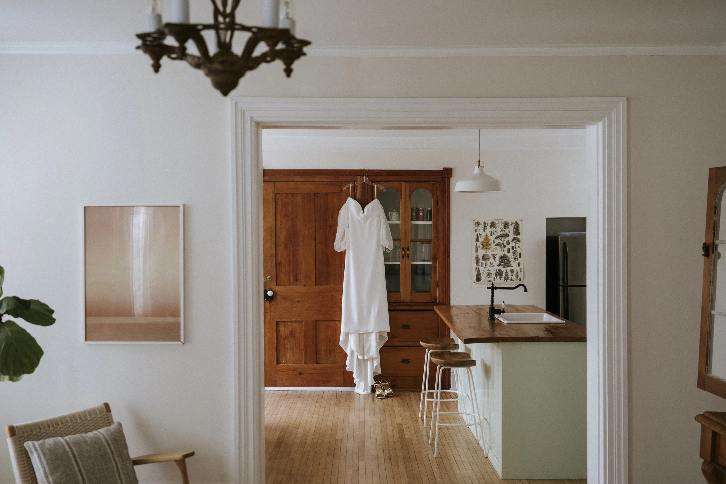 White wedding dress hanging on the doorway to a kitchen with wooden cabinets and bar stools.
