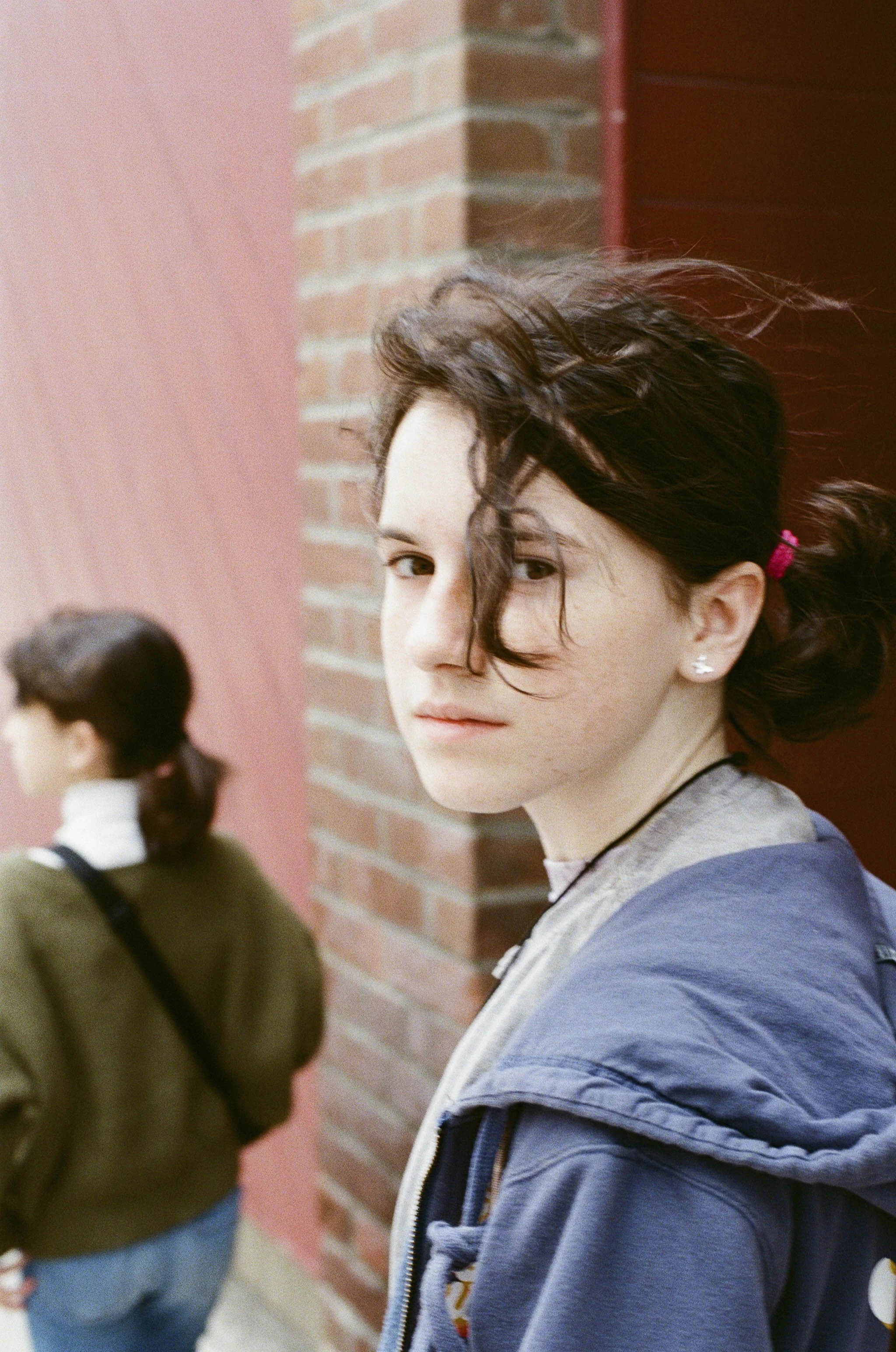 A young woman with dark hair tied in a bun, wearing earrings and a blue jacket, is standing near a brick wall, looking at the camera with a serious expression. A person with dark hair, wearing a green jacket and a black strap bag, is in the backgroun