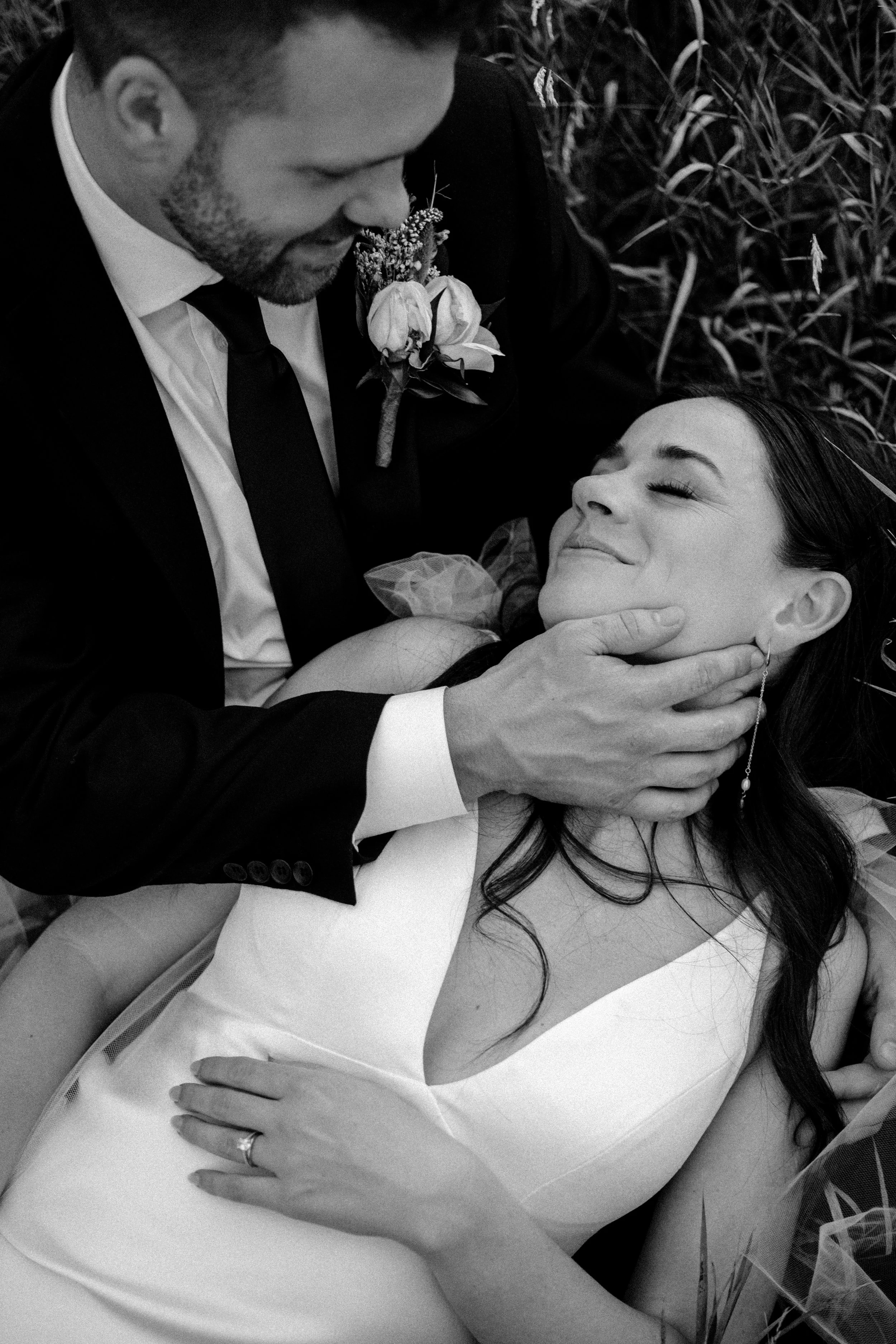 A black-and-white photo of a man in a suit holding a woman in a dress, lying on the grass during a wedding or romantic moment. The man gently touches the woman's face while she smiles peacefully.