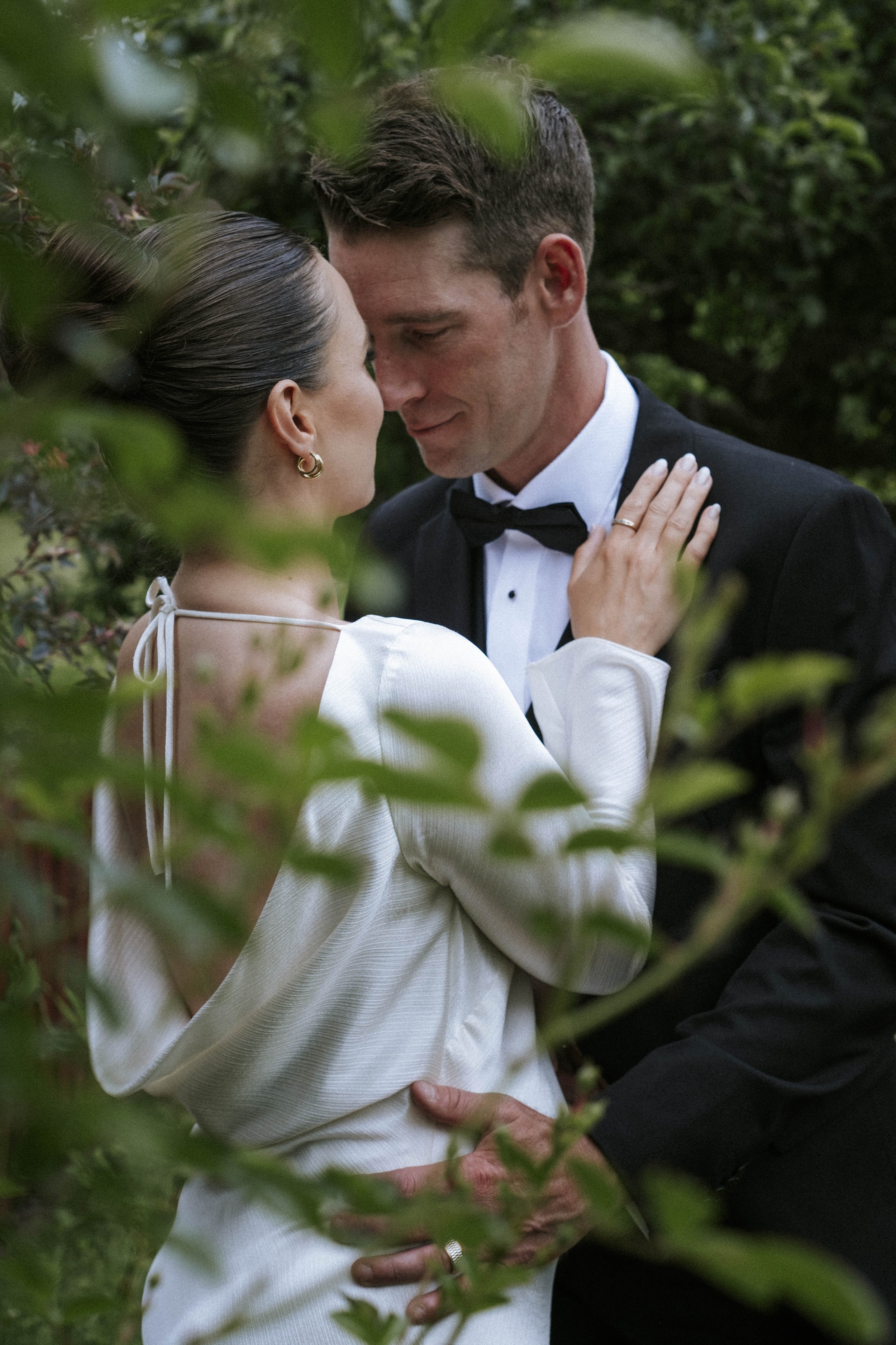 A couple dressed in formal wedding attire, embracing in a lush green outdoor setting, sharing a tender moment.