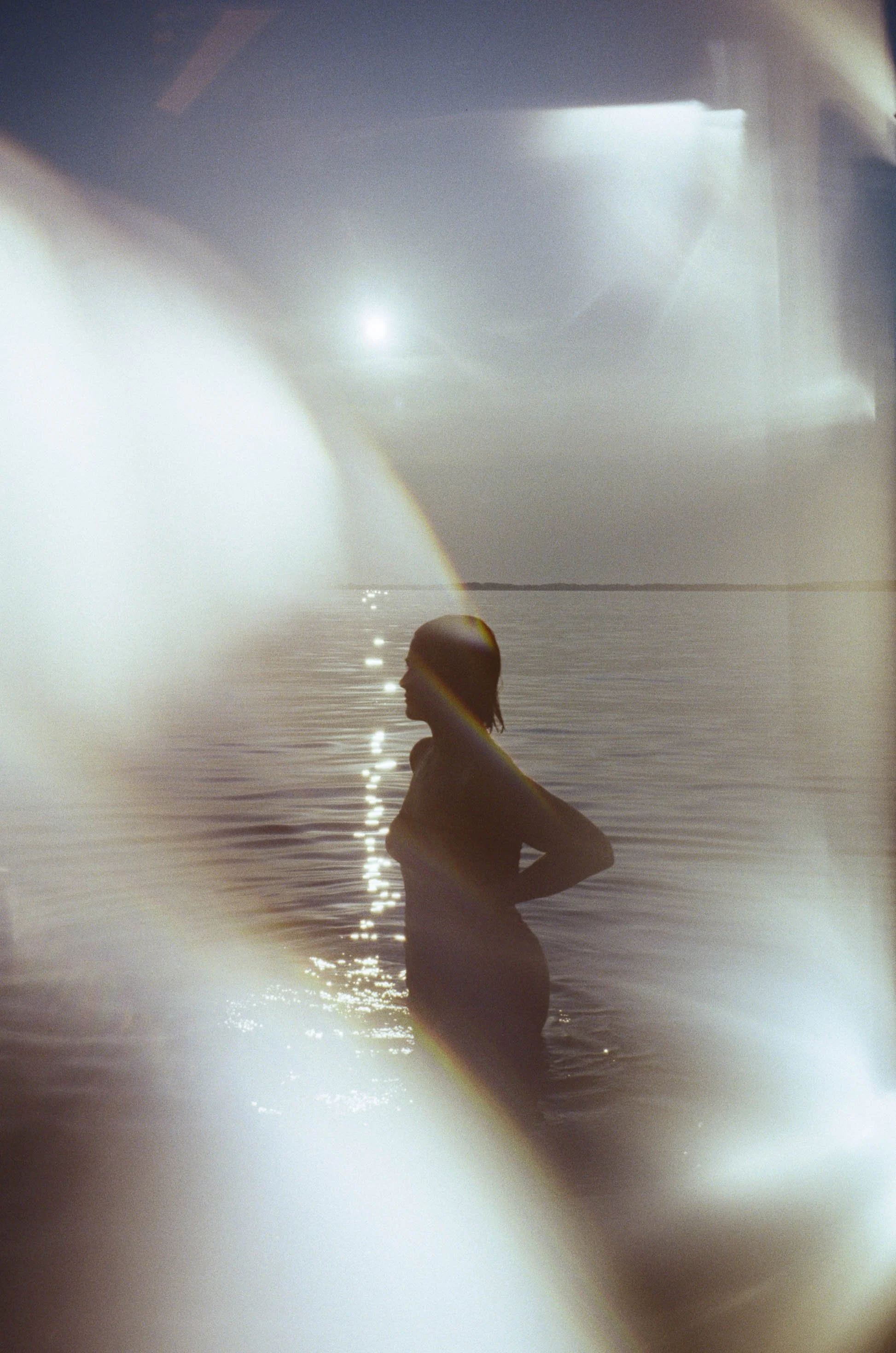 Silhouette of a woman standing in water at sunset with a rainbow reflection on her face and a lens flare effect.