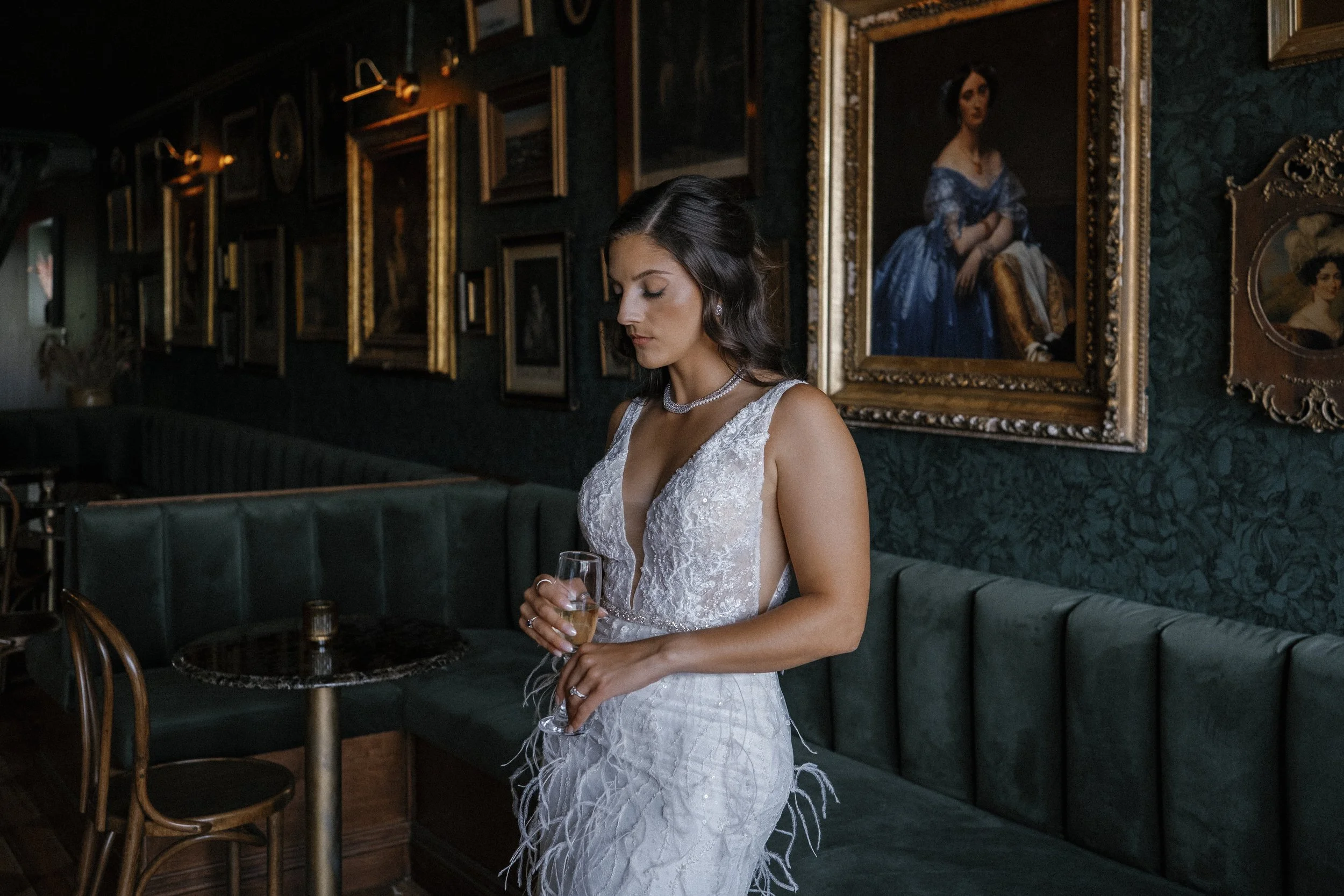 A woman in an elegant white lace dress holding a glass of champagne, standing in a richly decorated room with framed portraits on the dark green walls.