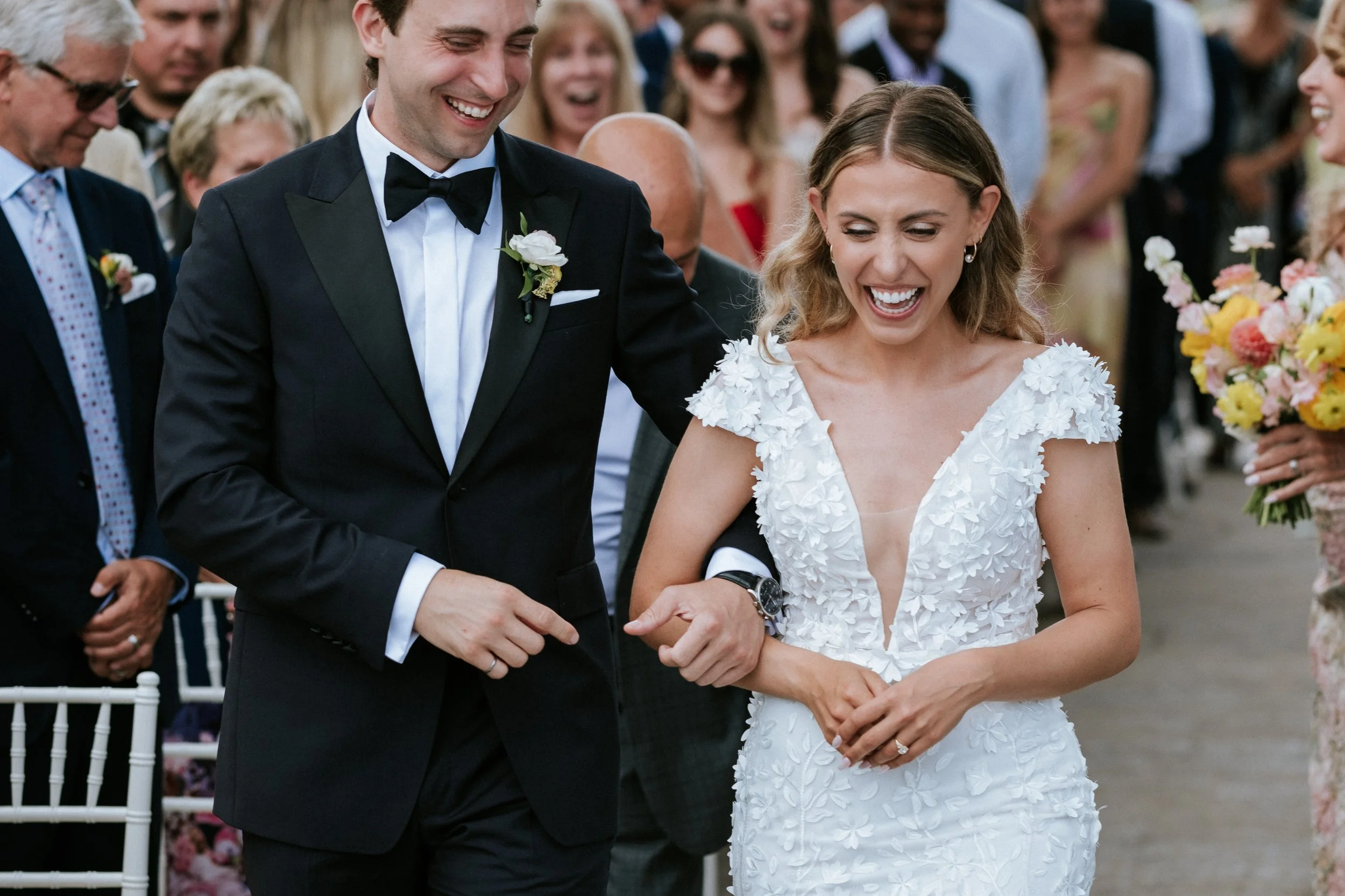 A happy bride and groom walking down the aisle, surrounded by guests. The bride is wearing a white wedding dress with floral details, and the groom is in a black tuxedo with a bow tie. Both are smiling and holding hands.