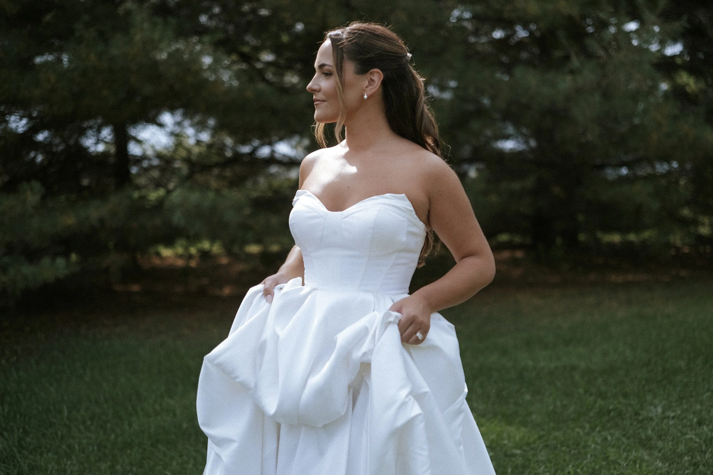 A woman in a strapless white wedding dress standing outdoors on grass with trees in the background.