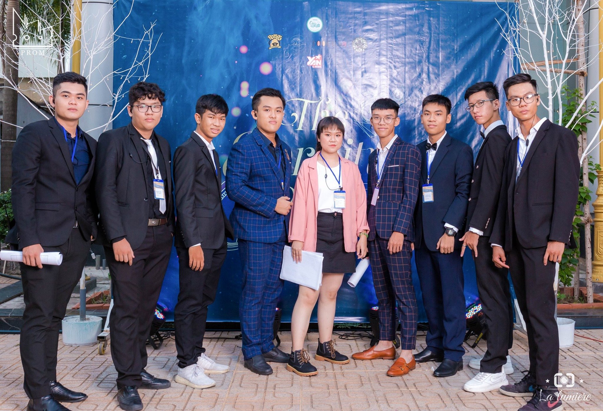 A group of nine young people dressed in formal business attire, standing outdoors in front of a blue backdrop with text. They are posing for a photo, and some are holding rolled-up papers or folders.