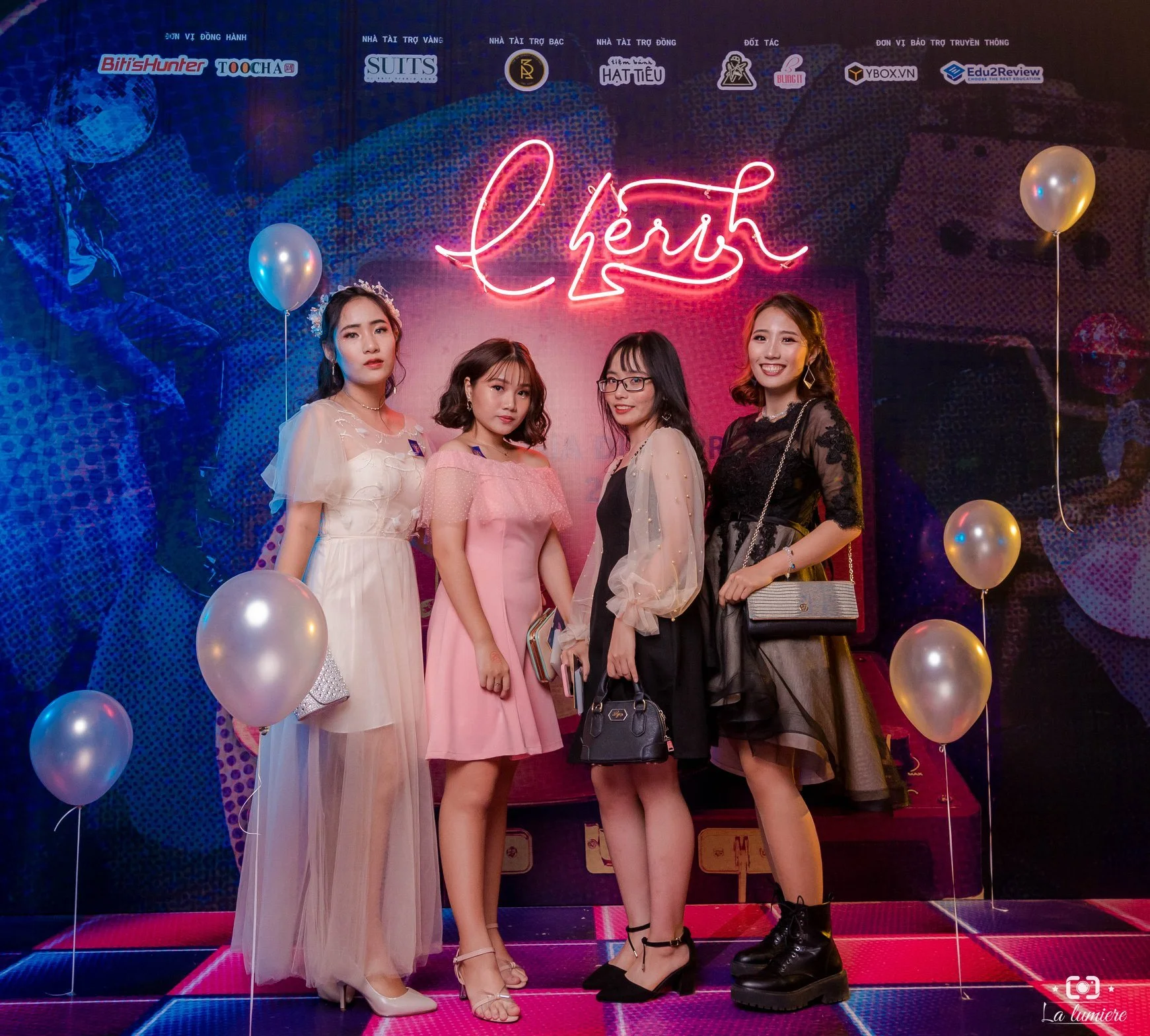 Four young women are standing together on a colorful stage with balloons, in front of a neon sign that reads 'cherk'. They are dressed stylishly in dresses and accessories, with a backdrop featuring a large image and various sponsor logos at the top.