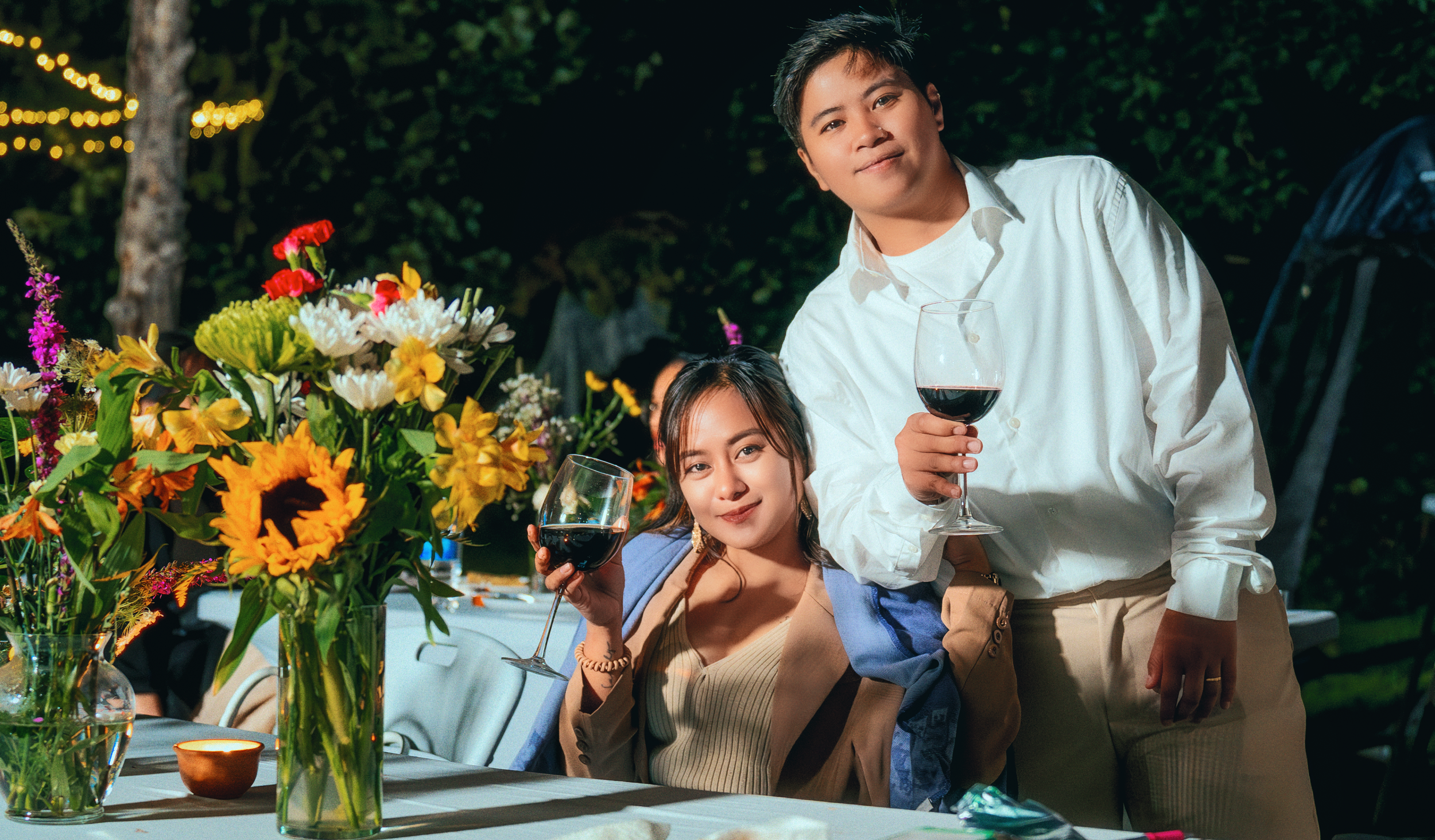A young woman and young man celebrating with red wine at an outdoor evening party, seated at a table decorated with colorful flowers, with trees and string lights in the background.
