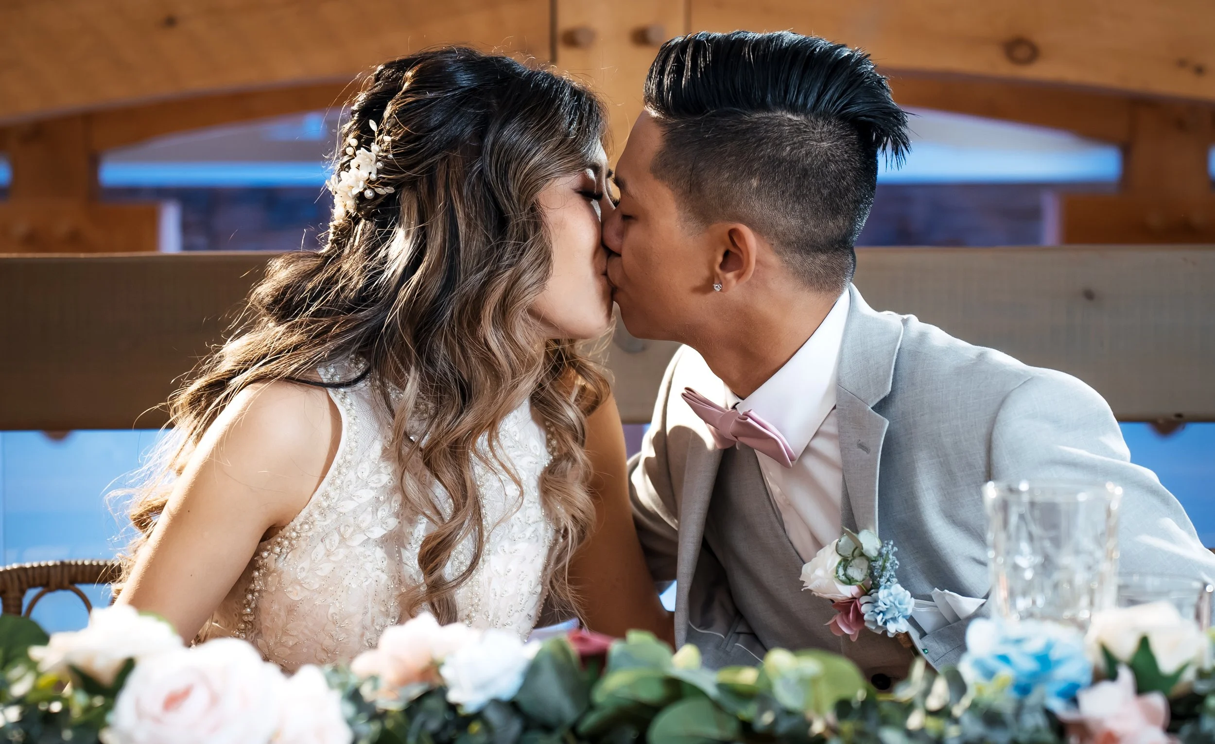 A couple kisses at their wedding reception, with flowers and glasses on the table, and a wooden arch in the background.