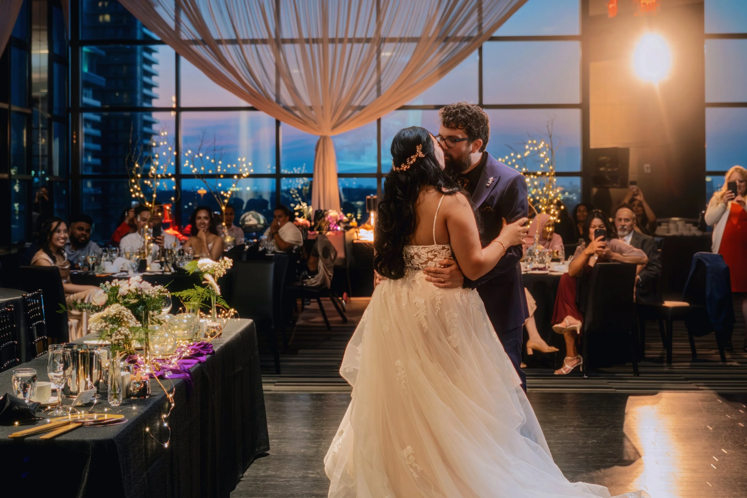 A bride and groom kissing while dancing at their wedding reception, with guests seated at decorated tables in the background.