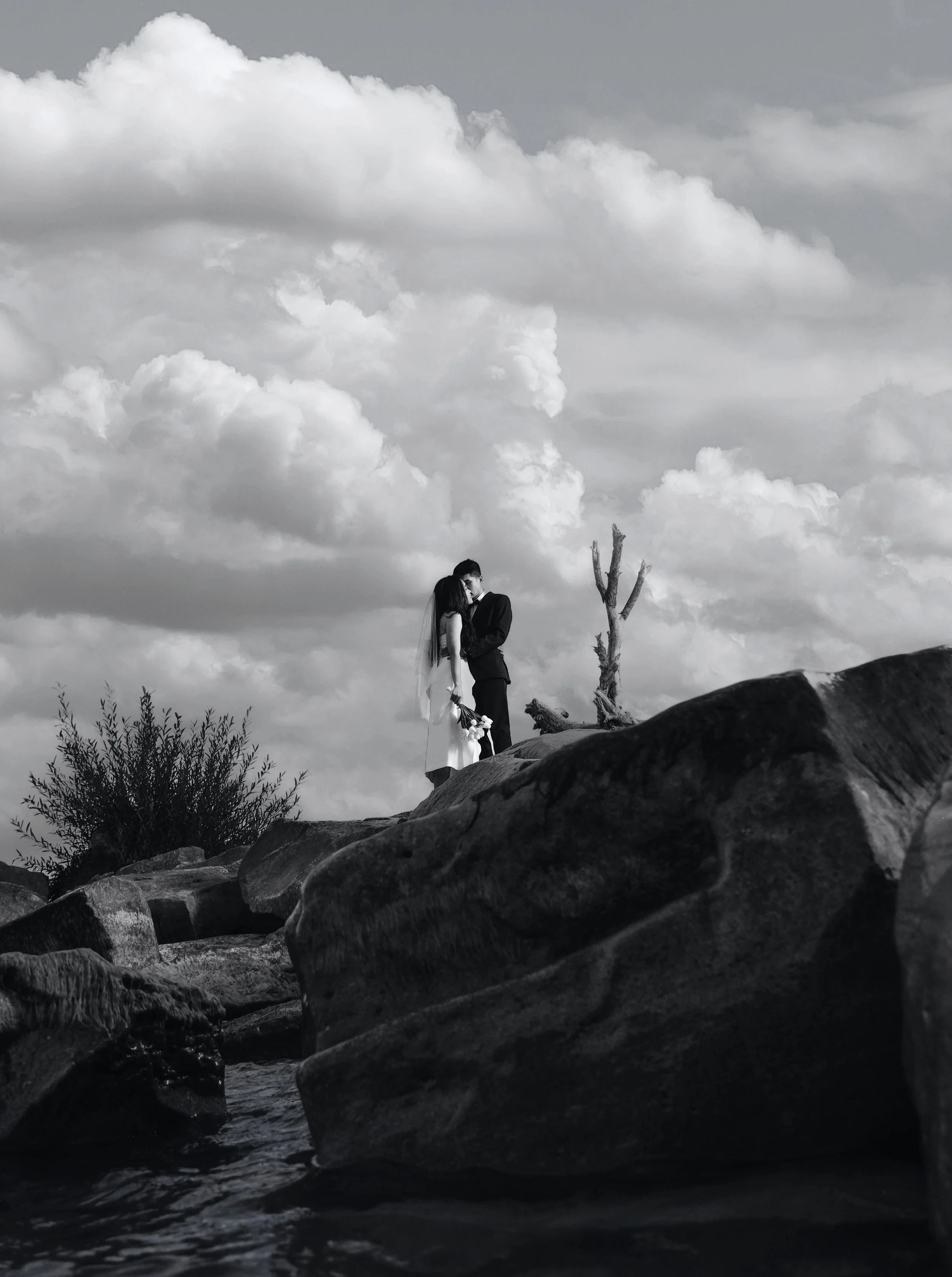 A black and white photo of a couple standing on rocks by a body of water, embracing each other, with a cloudy sky in the background.