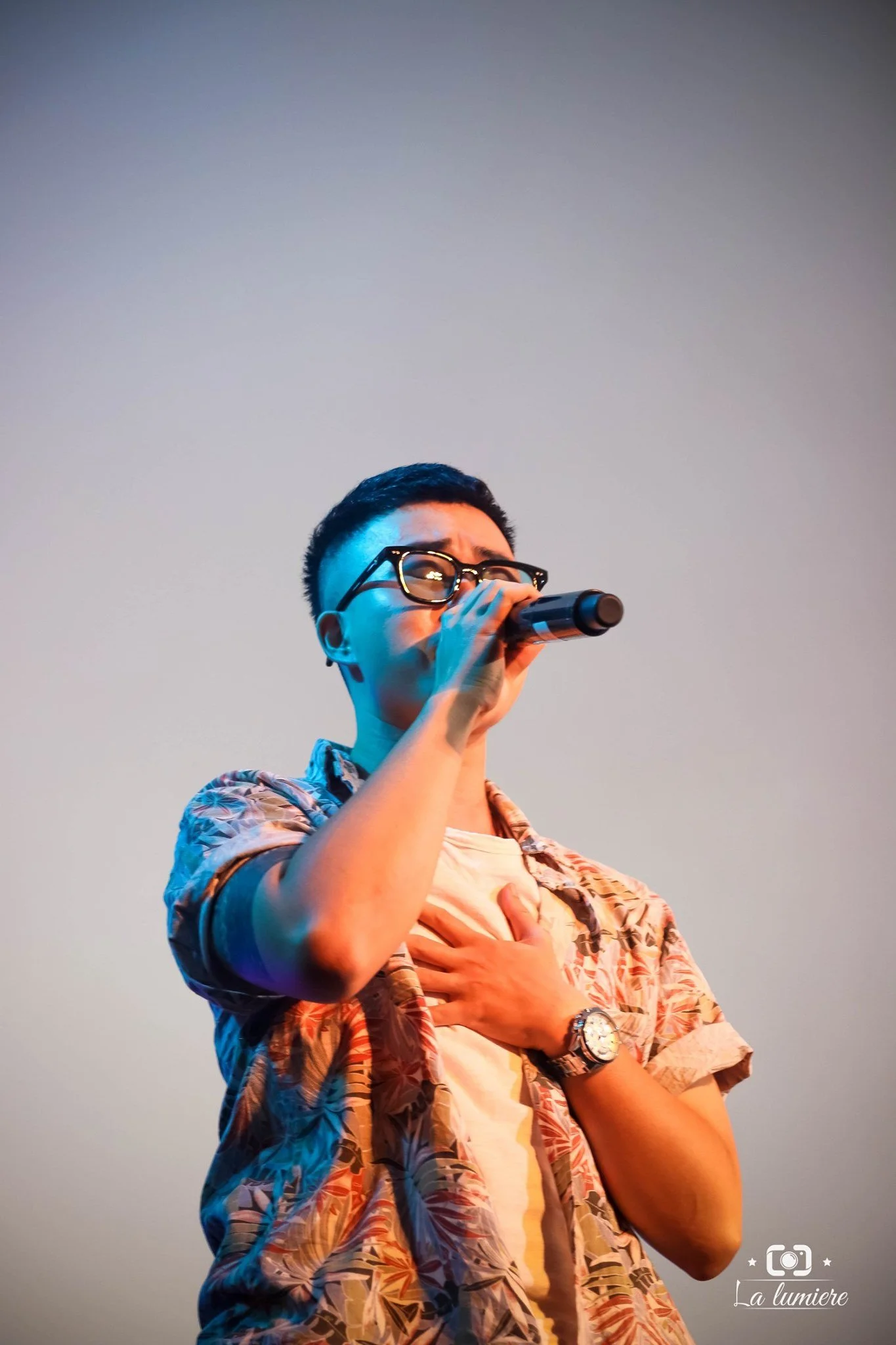 A young man singing into a microphone on stage, wearing glasses, a floral shirt, and a watch, with his hand on his chest.